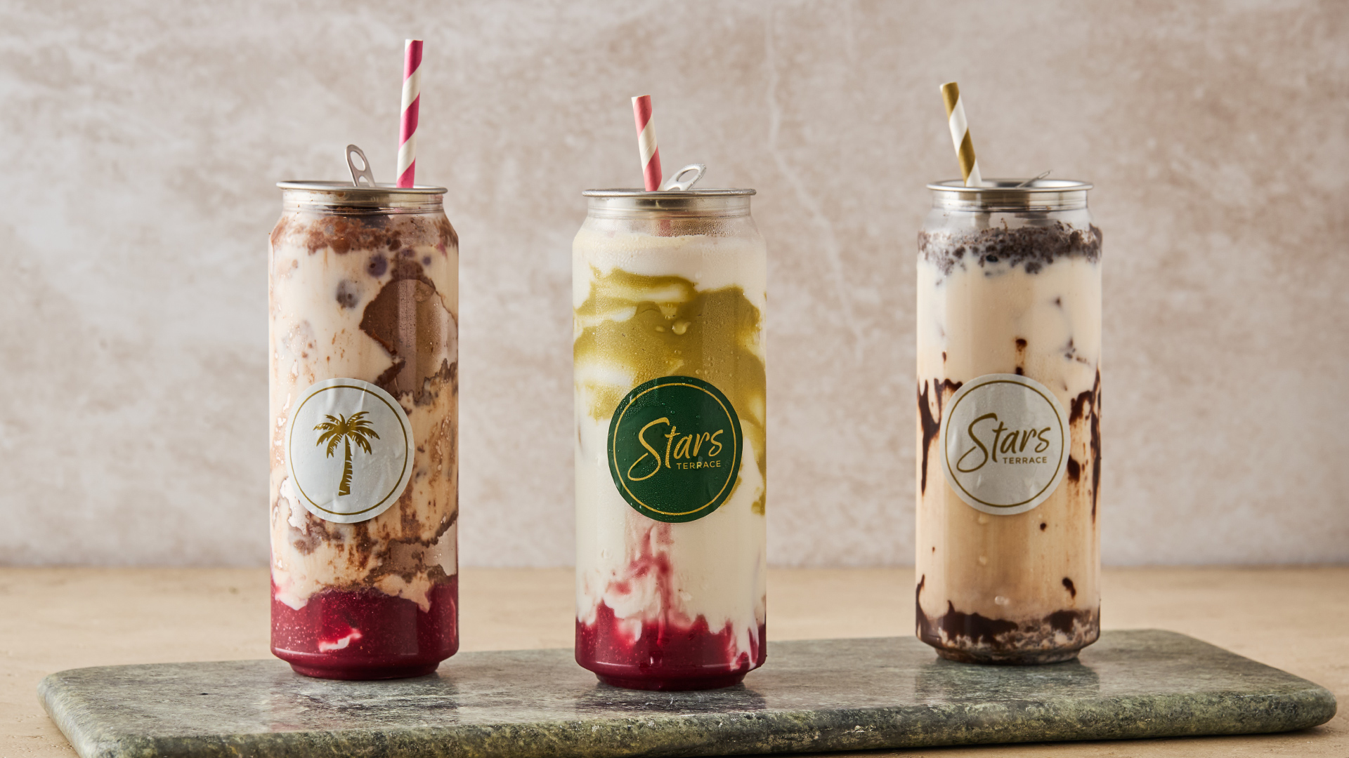 Three tall glass cups filled with layered milkshakes in different flavors, topped with whipped cream and accompanied by colorful striped straws, are arranged on a stone tray against a neutral background.