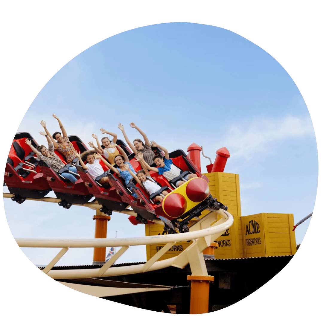 People riding a roller coaster at Warner Bros. Movie World with arms raised, against a clear blue sky.
