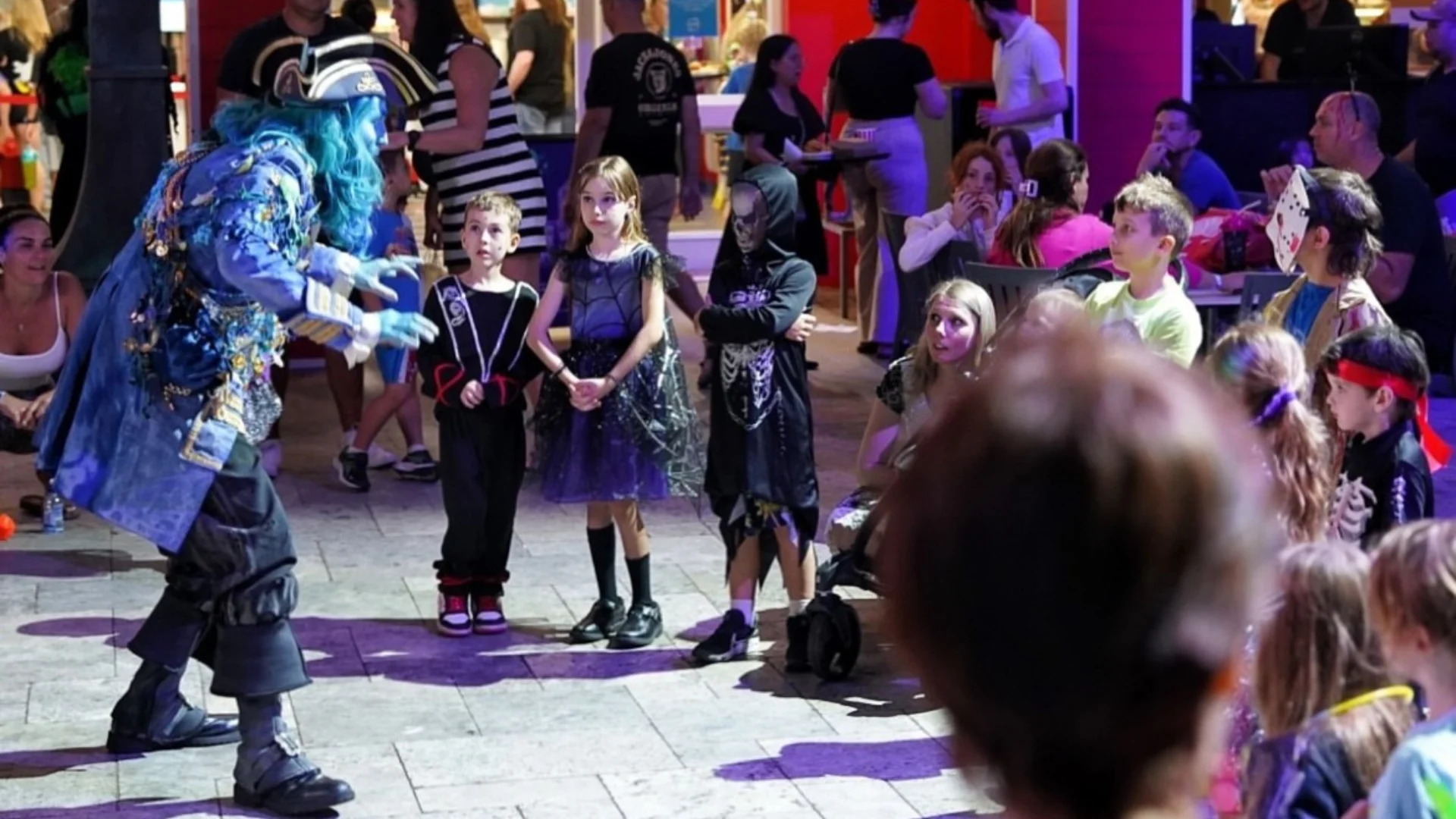 A performer in a blue costume entertains a group of children dressed in Halloween costumes, while adults watch from tables in the background. The scene is lively and colorful, with a festive atmosphere.