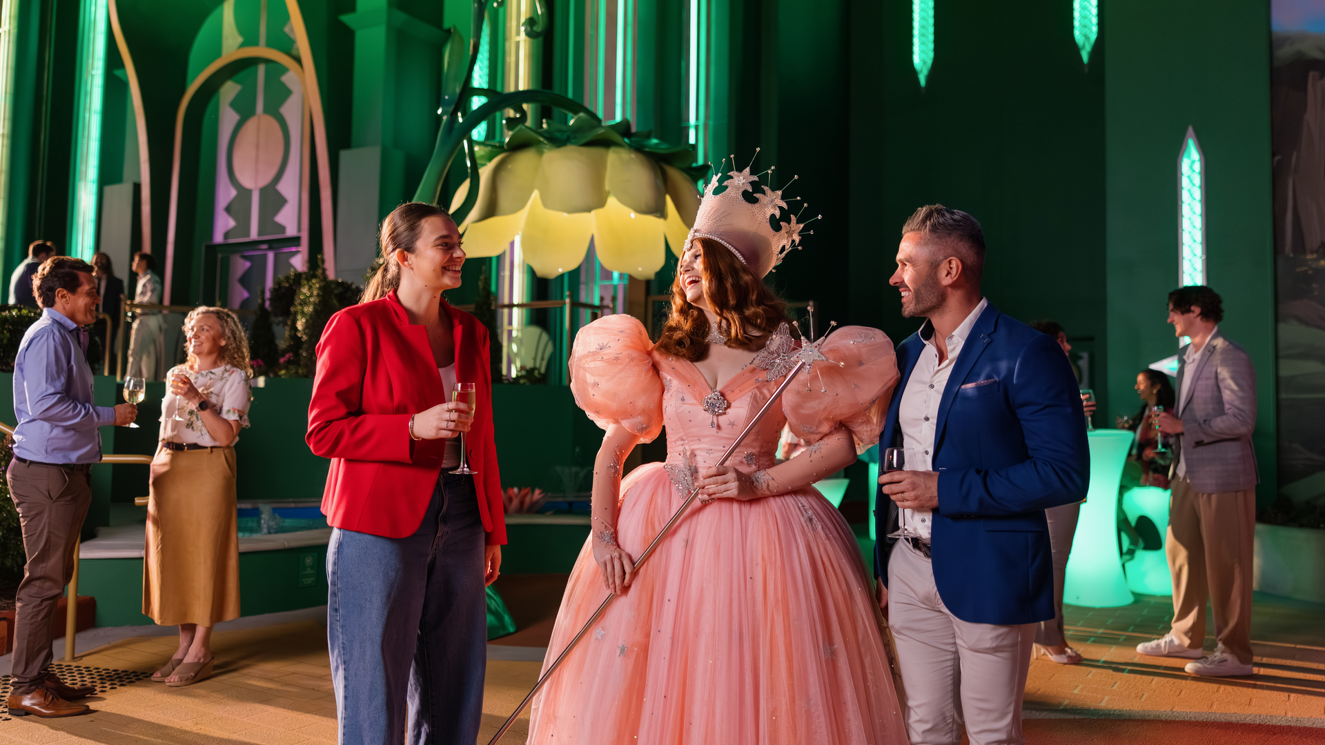 A woman in a pink ball gown and crown holds a wand, talking to a smiling man and woman at a lively party in a colorful, fantasy-themed setting with green and gold decor.