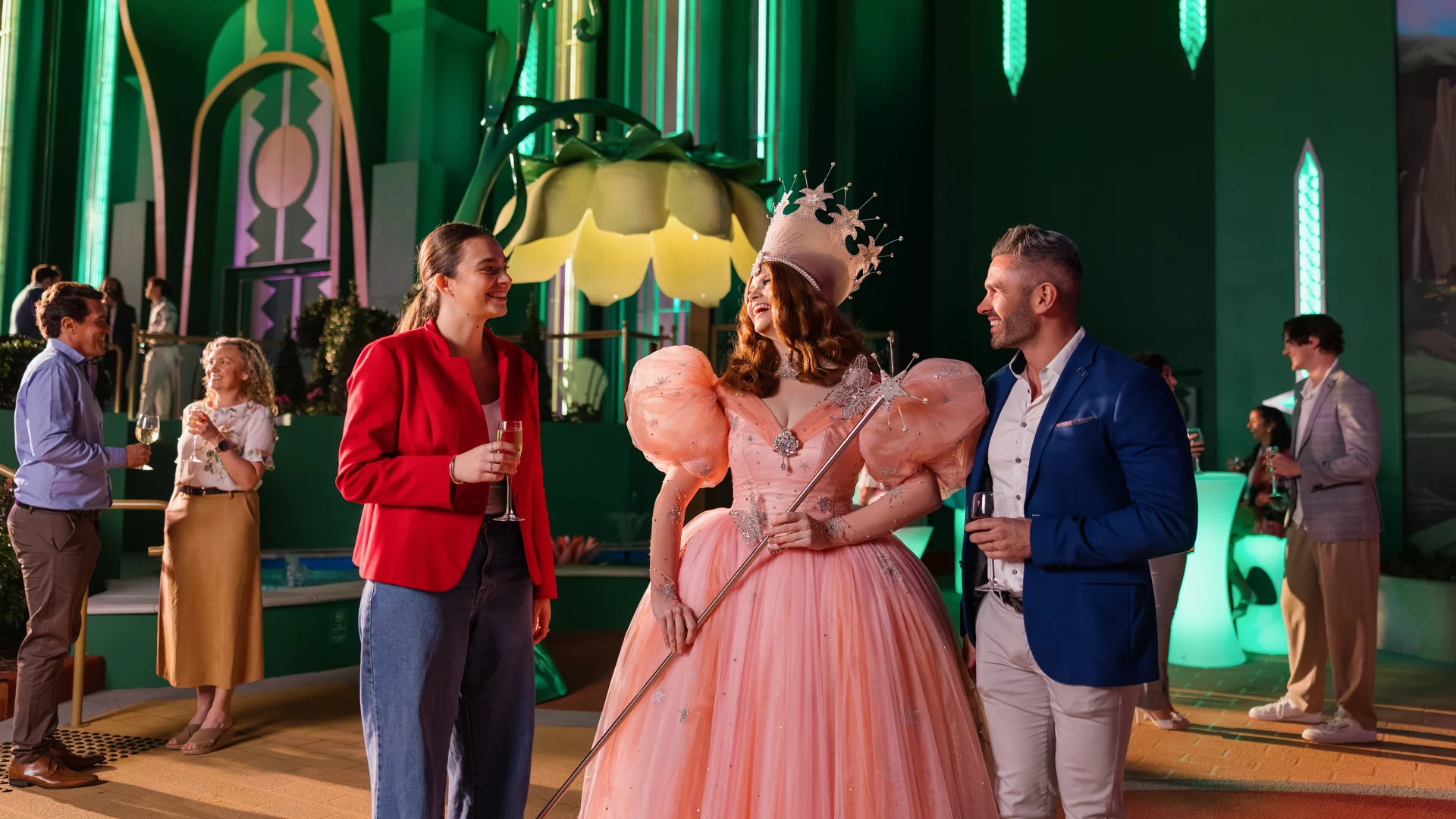 A woman in a pink ball gown and crown holds a wand, talking to a smiling man and woman at a lively party in a colorful, fantasy-themed setting with green and gold decor.