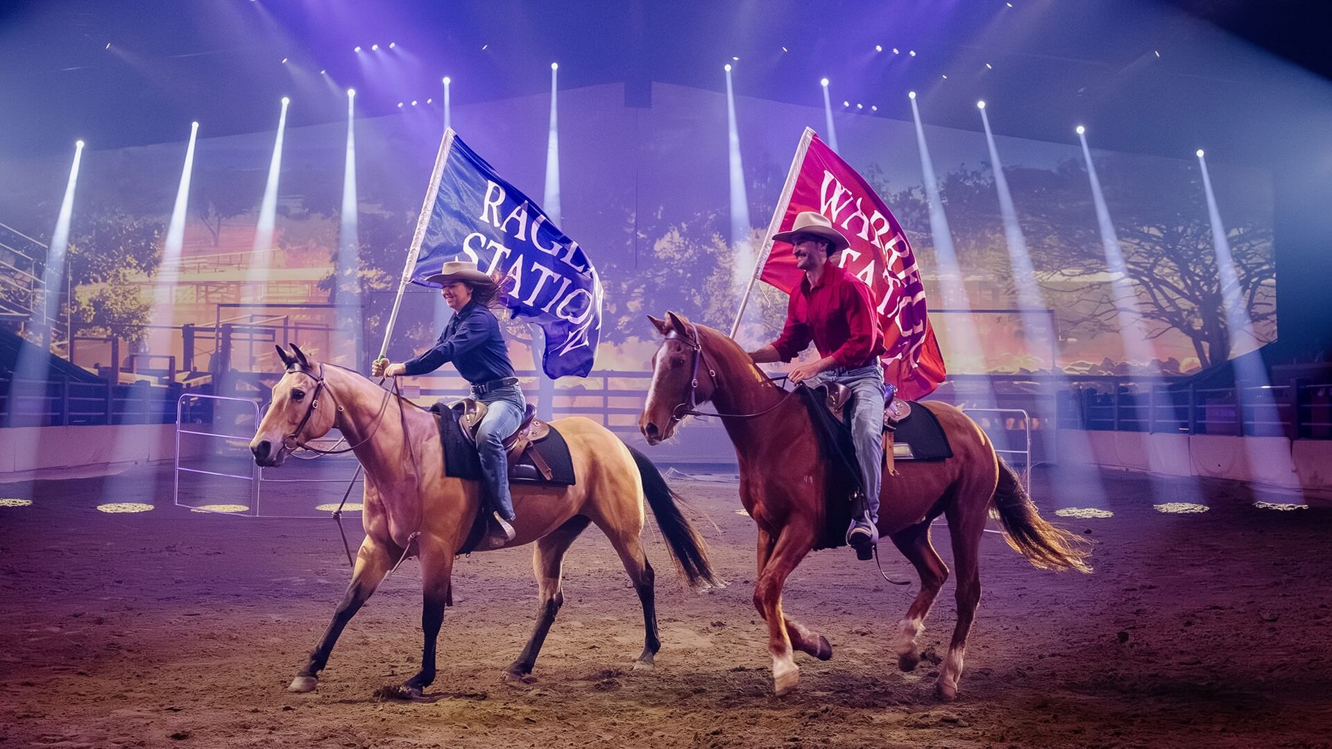 Two horseback riders perform in an indoor arena under dramatic stage lighting, each carrying large station-branded flags during an Australian outback-themed live show.