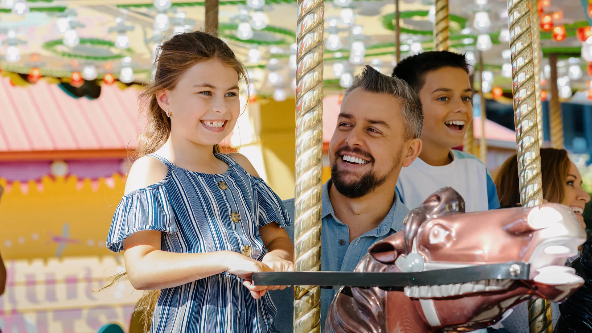 A young girl and an adult man smile while riding a carousel together; other children are visible in the background.
