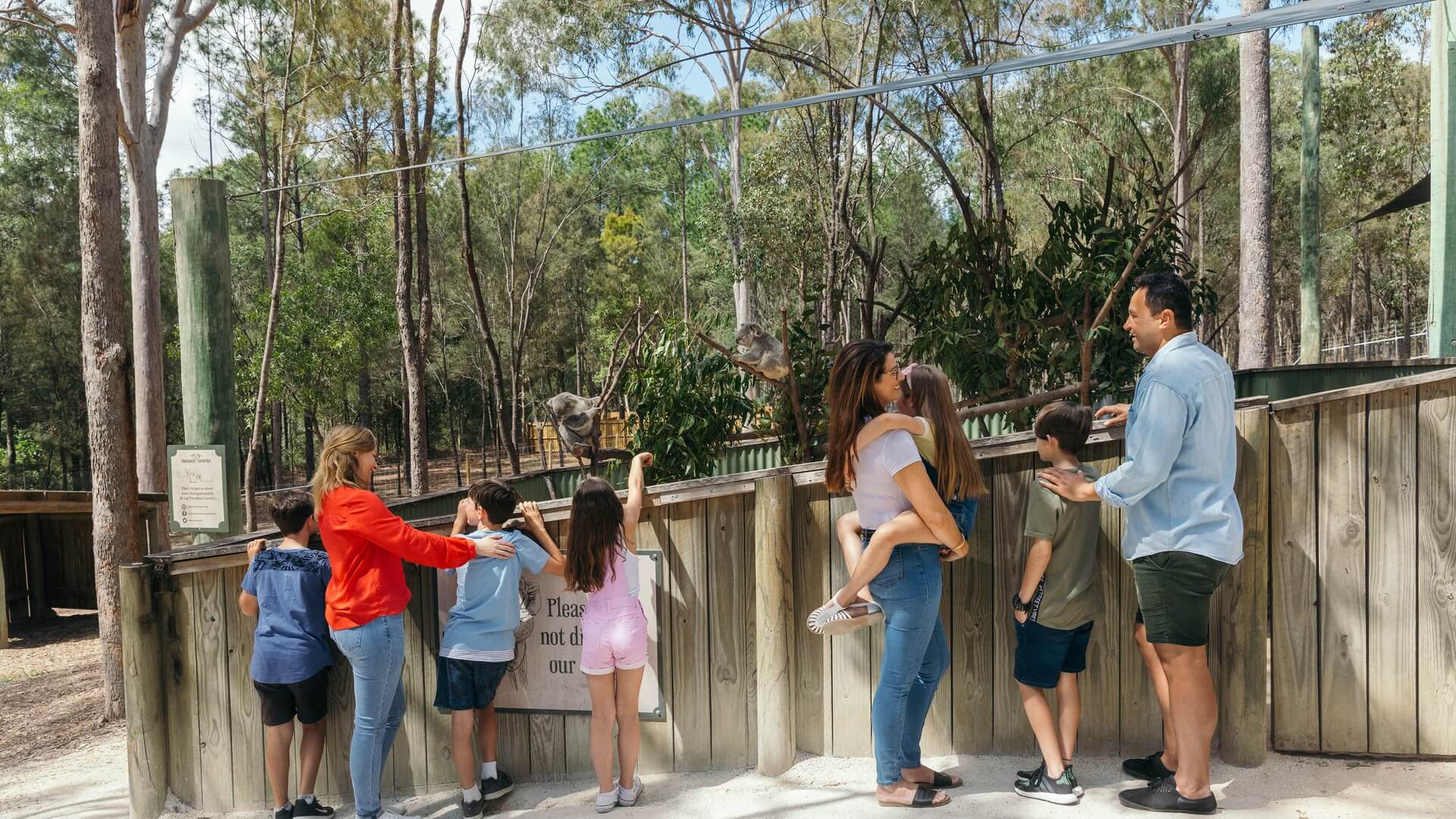 A group of adults and children stand at a wooden fence in an outdoor zoo, observing animals in an enclosure surrounded by trees.