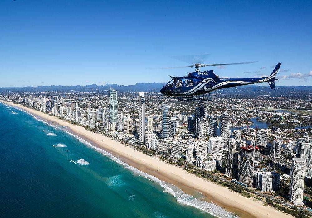 A helicopter flies over a coastal city with high-rise buildings, a sandy beach, and a blue ocean under a clear blue sky.