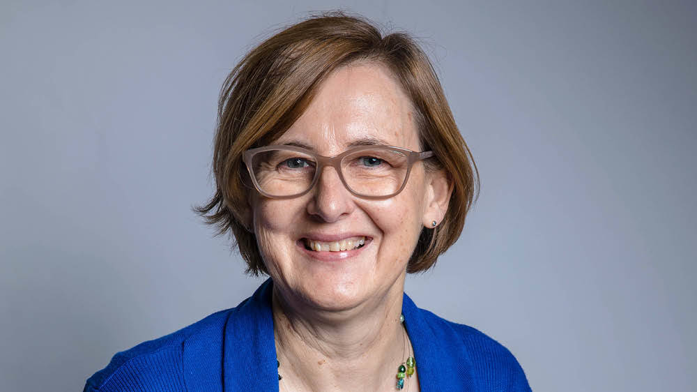A middle-aged woman with short light brown hair, glasses, and a blue top smiles at the camera against a plain background.