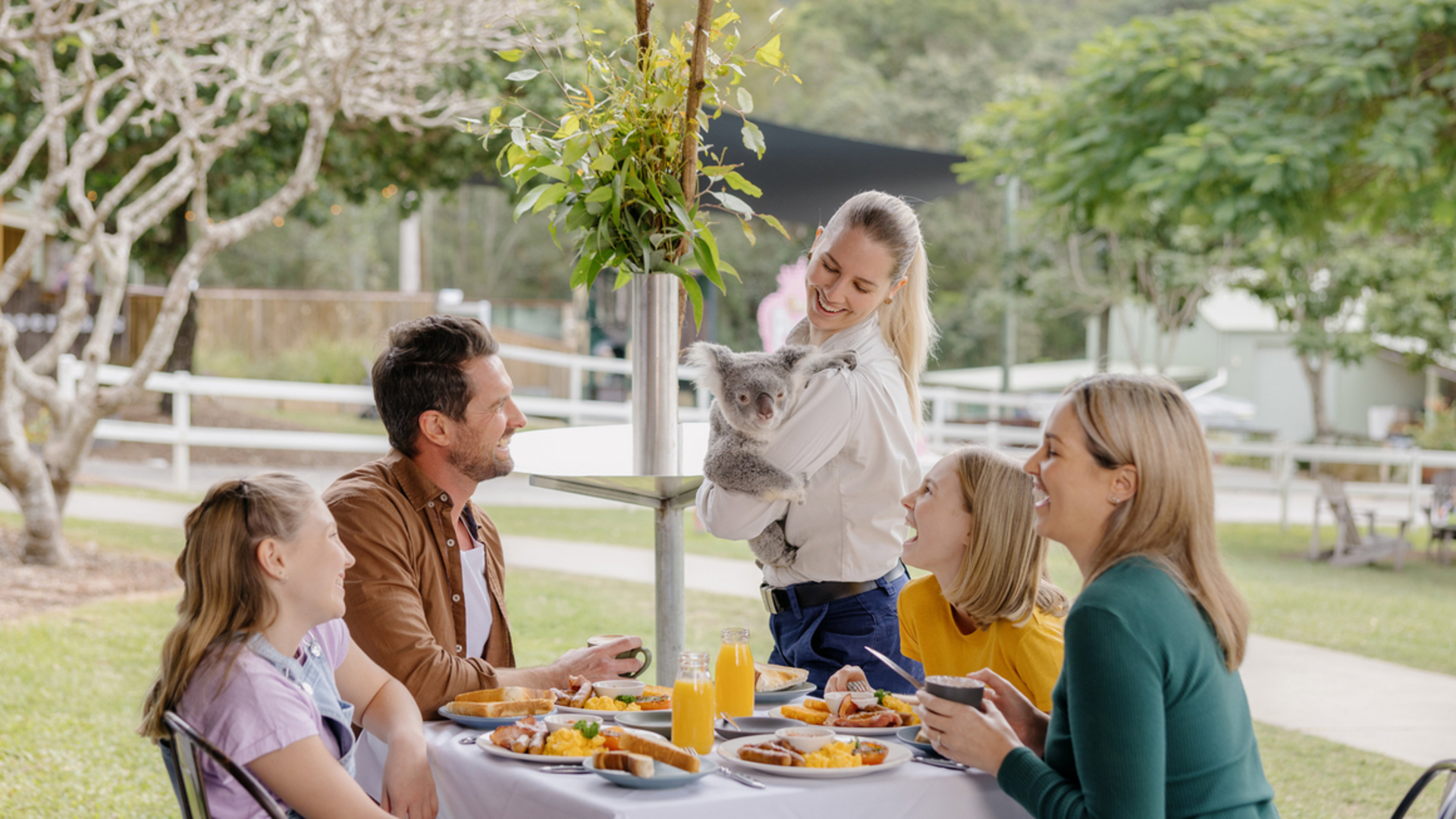 A group sitting at an outdoor table with breakfast food and drinks. A woman is holding a koala, engaging with the others. Trees and a pathway are visible in the background.