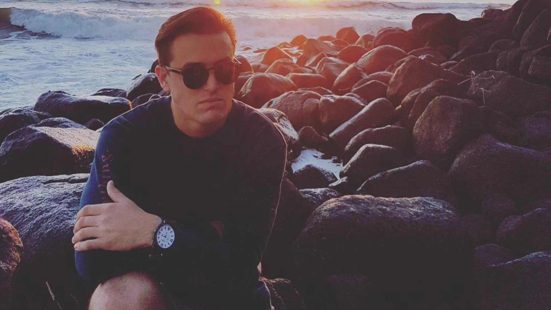 A person wearing dark sunglasses and a watch sits on large rocks near the ocean at sunset, with waves and sunlight visible in the background.