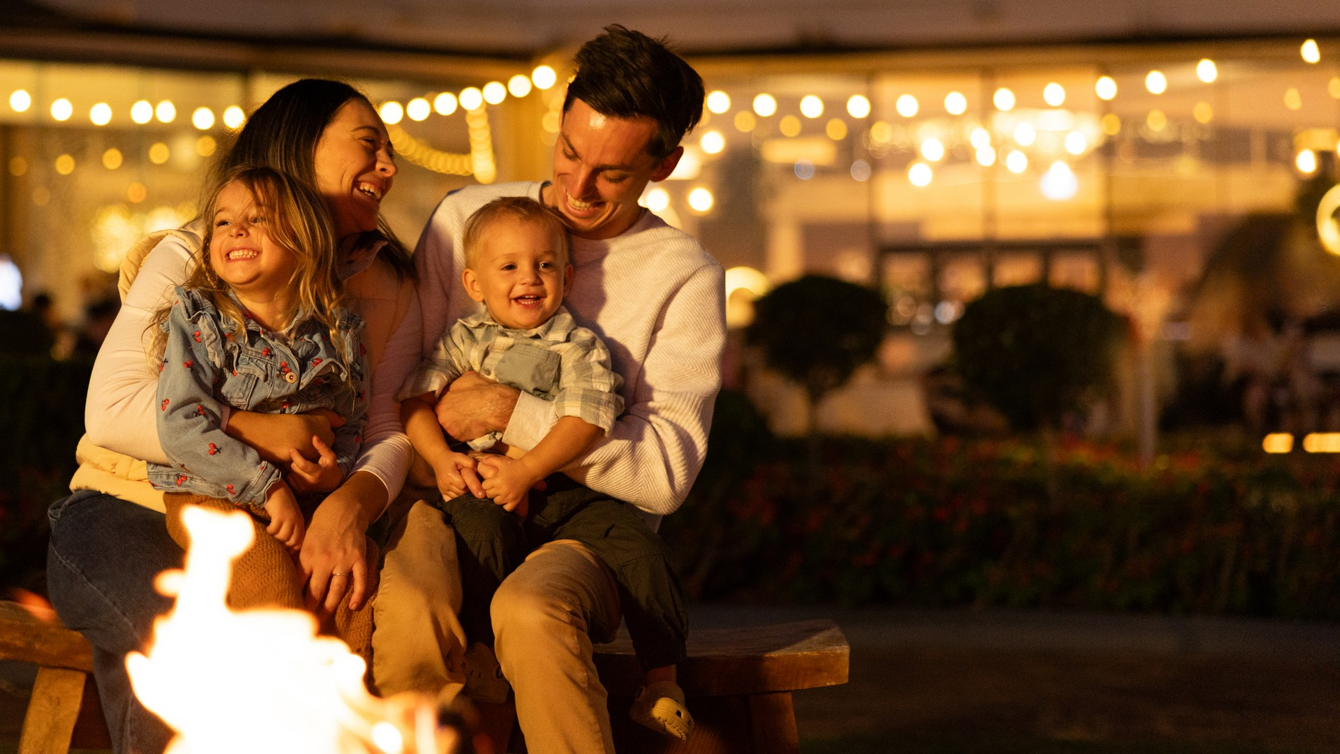 A smiling family of four sits together by a firepit at night, sharing laughter. String lights and blurred greenery create a warm, festive background.