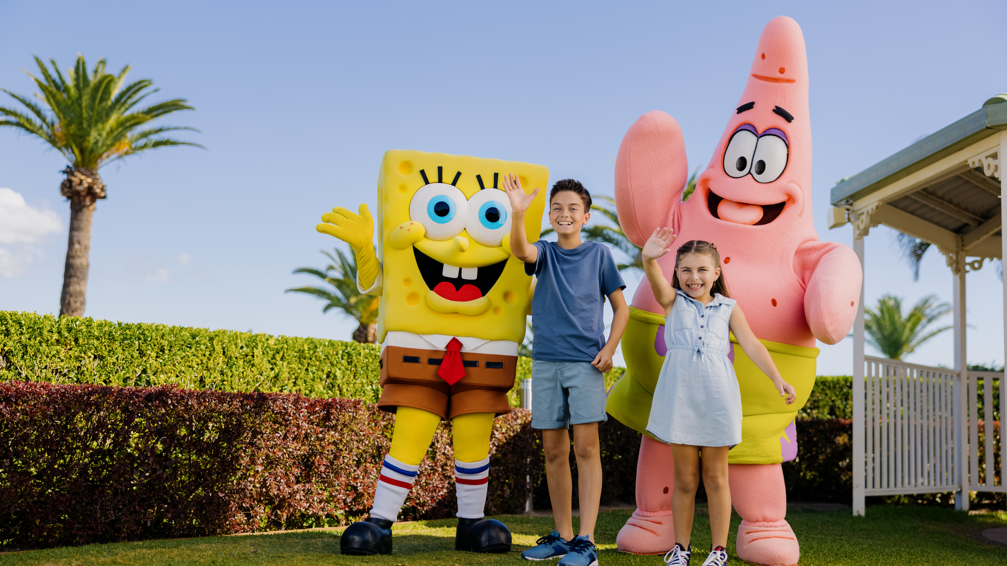 Two children stand next to costumed characters resembling a cartoon sponge and starfish, outside near greenery and a gazebo.