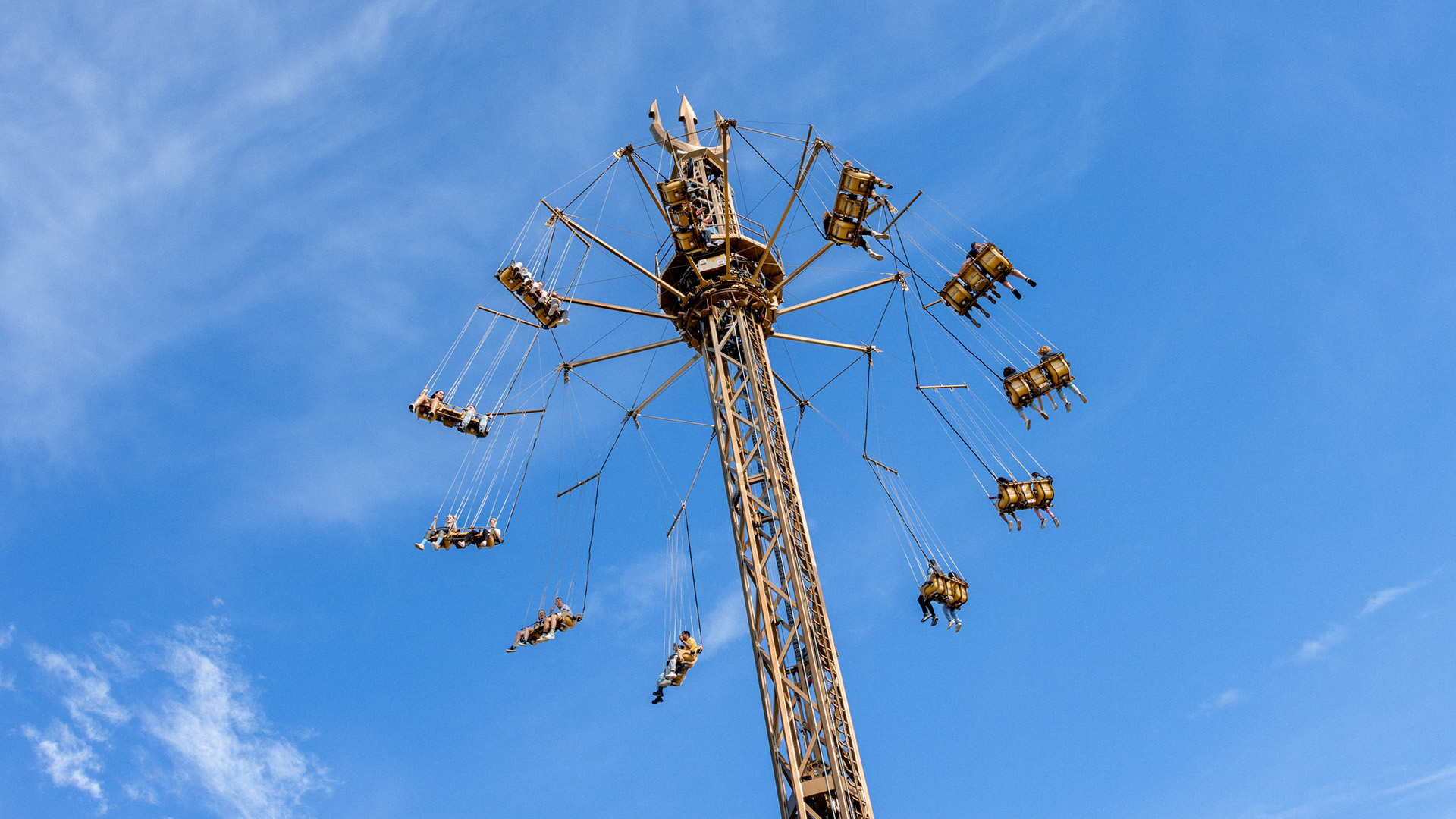 People are seated in swings attached to a tall amusement park ride, spinning in the air against a blue sky with wispy clouds.