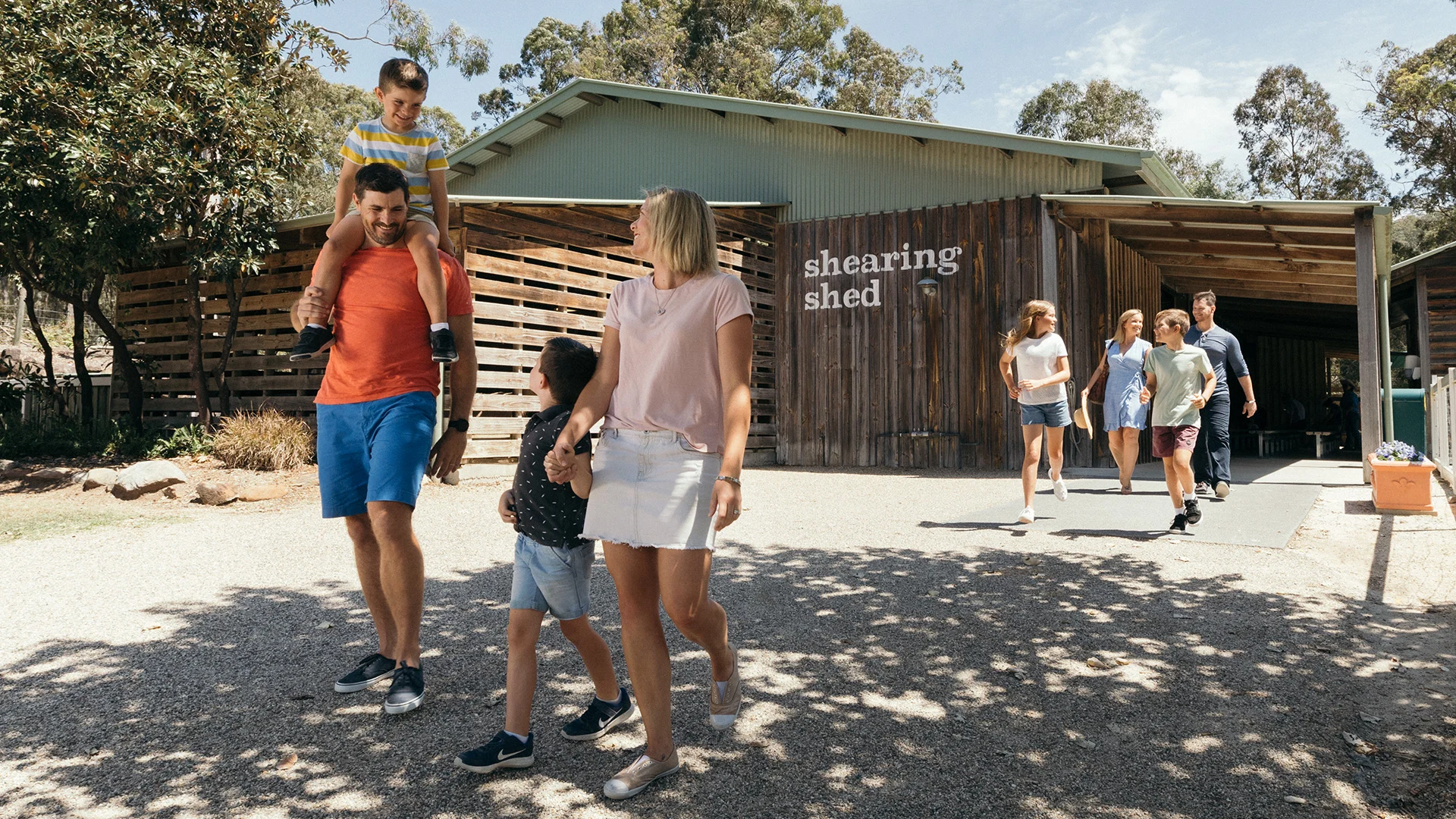A family with two children walks outside a building labeled "shearing shed" at Paradise Country, while two other people walk behind them under a clear sky.