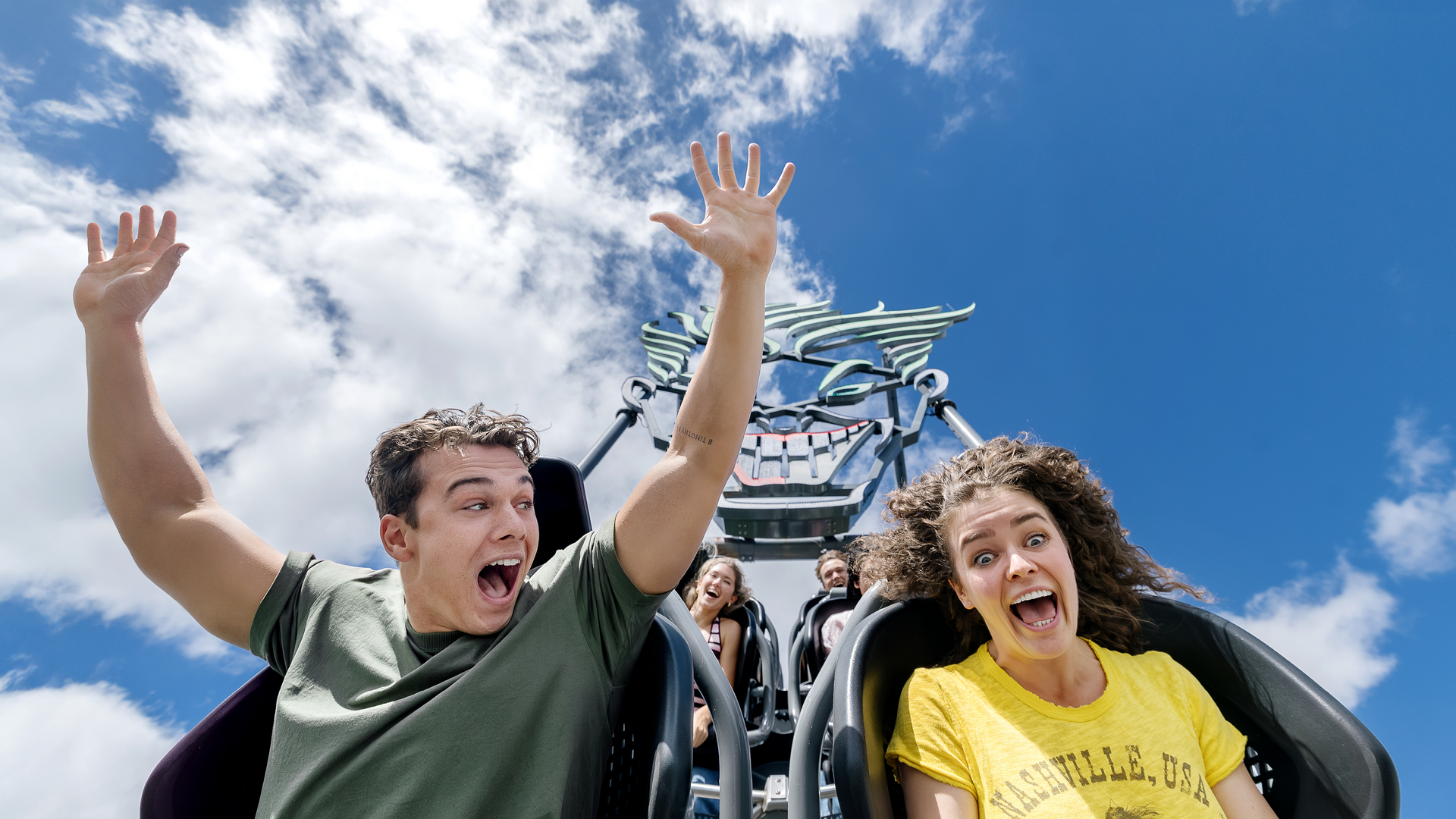 Two people with excited expressions ride a roller coaster with their hands up against a bright blue sky with clouds, while others ride behind them.