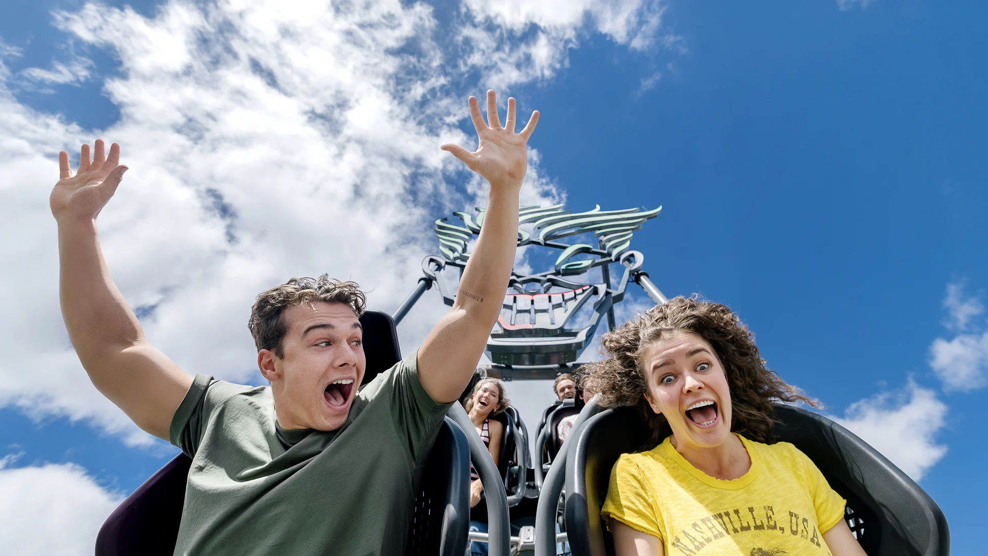 Two people with excited expressions ride a roller coaster with their hands up against a bright blue sky with clouds, while others ride behind them.