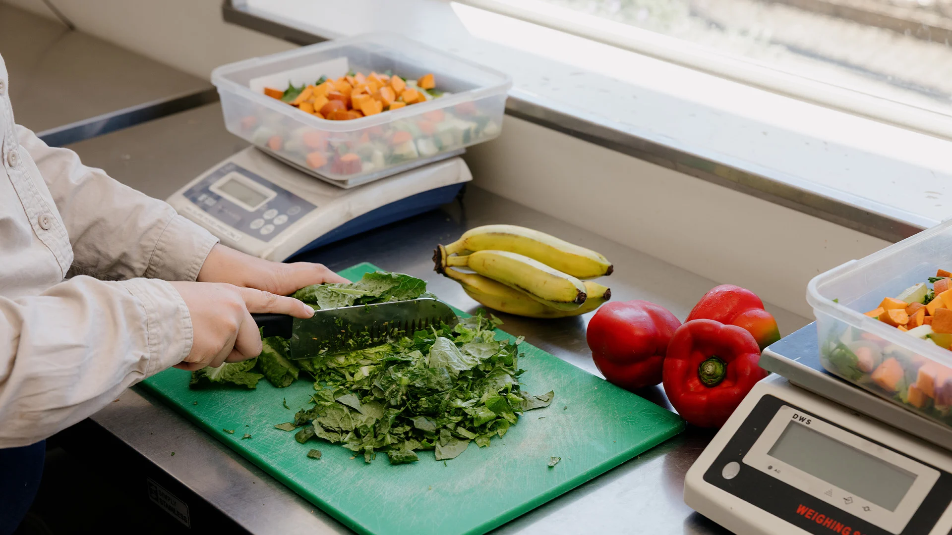 A person chops leafy greens on a green cutting board beside red bell peppers, bananas, and containers of diced produce in a kitchen setting. Digital scales and a food analyzer are also visible on the counter.