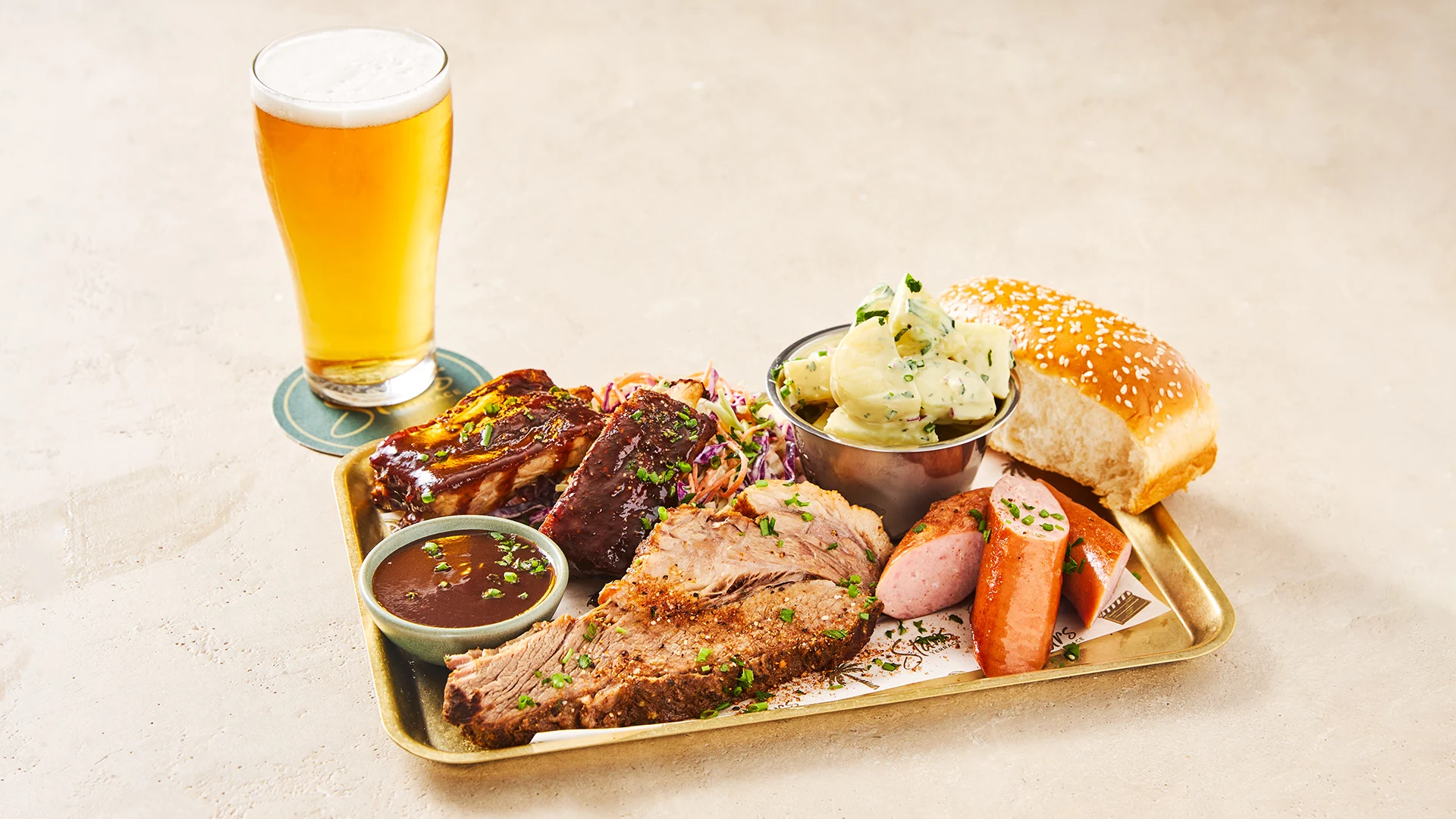 A tray with barbecue brisket, ribs, sausage, potato salad, barbecue sauce, coleslaw, and a sesame seed bun is next to a glass of beer on a coaster, set on a light-colored surface.