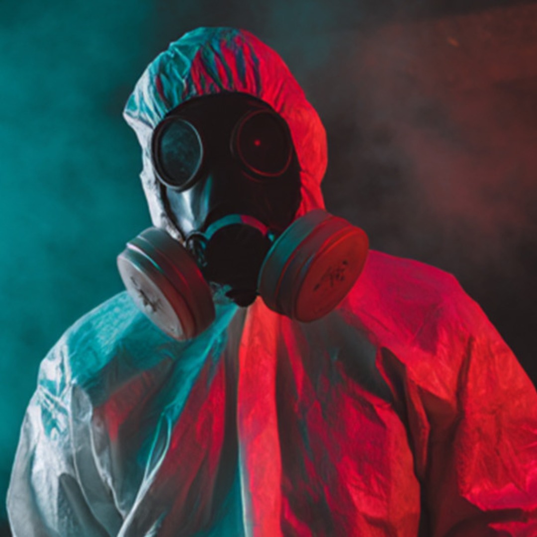 A person wearing a hazmat suit and a gas mask stands in dramatic lighting with blue and red tones, surrounded by mist or smoke.