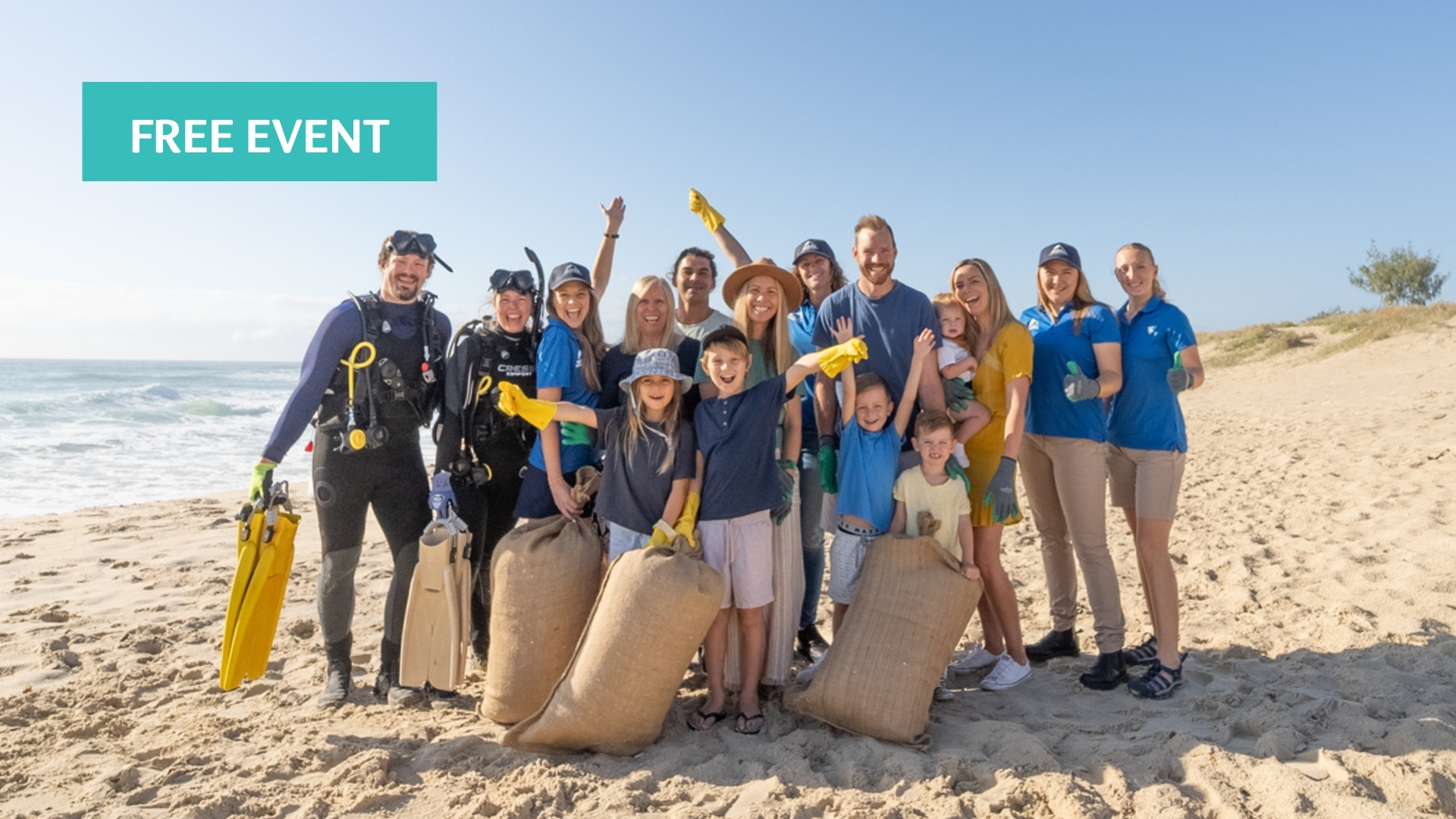 A diverse group of volunteers, including adults, kids, and scuba divers, smile and pose with large bags of collected trash on a sunny beach. A “Free Event” label is in the top left corner.