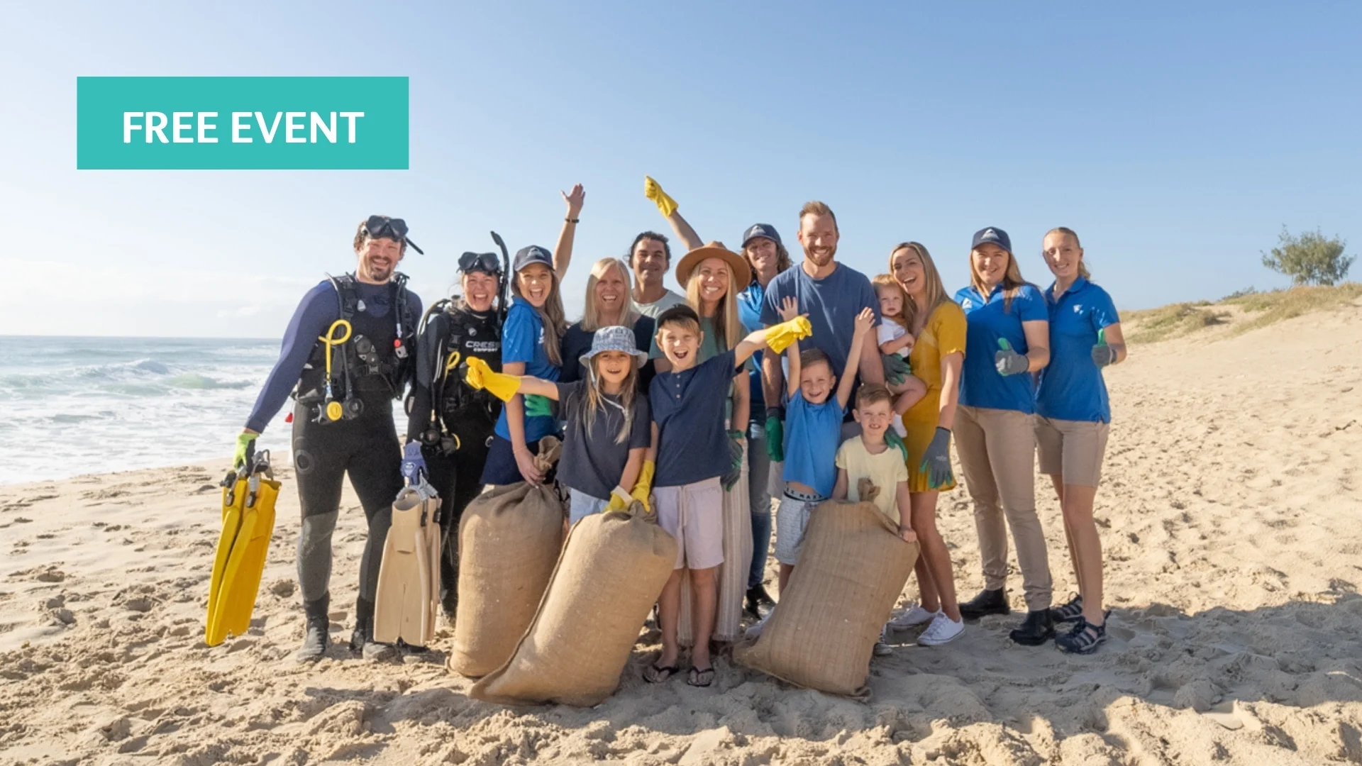 A diverse group of volunteers, including adults, kids, and scuba divers, smile and pose with large bags of collected trash on a sunny beach. A “Free Event” label is in the top left corner.
