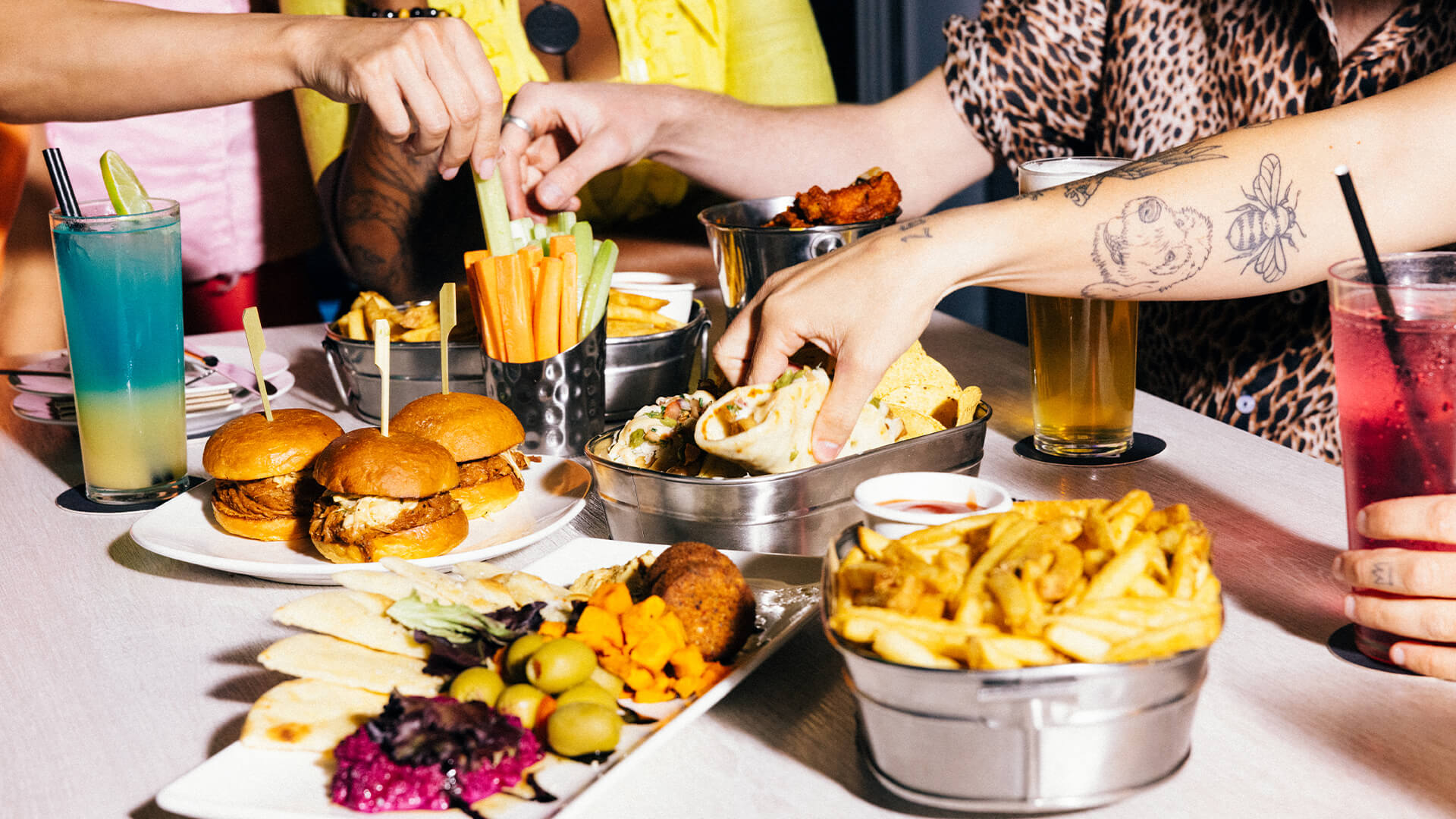 People enjoying a meal at a table with sliders, fries, vegetables, wraps, and drinks. Hands reaching for food.