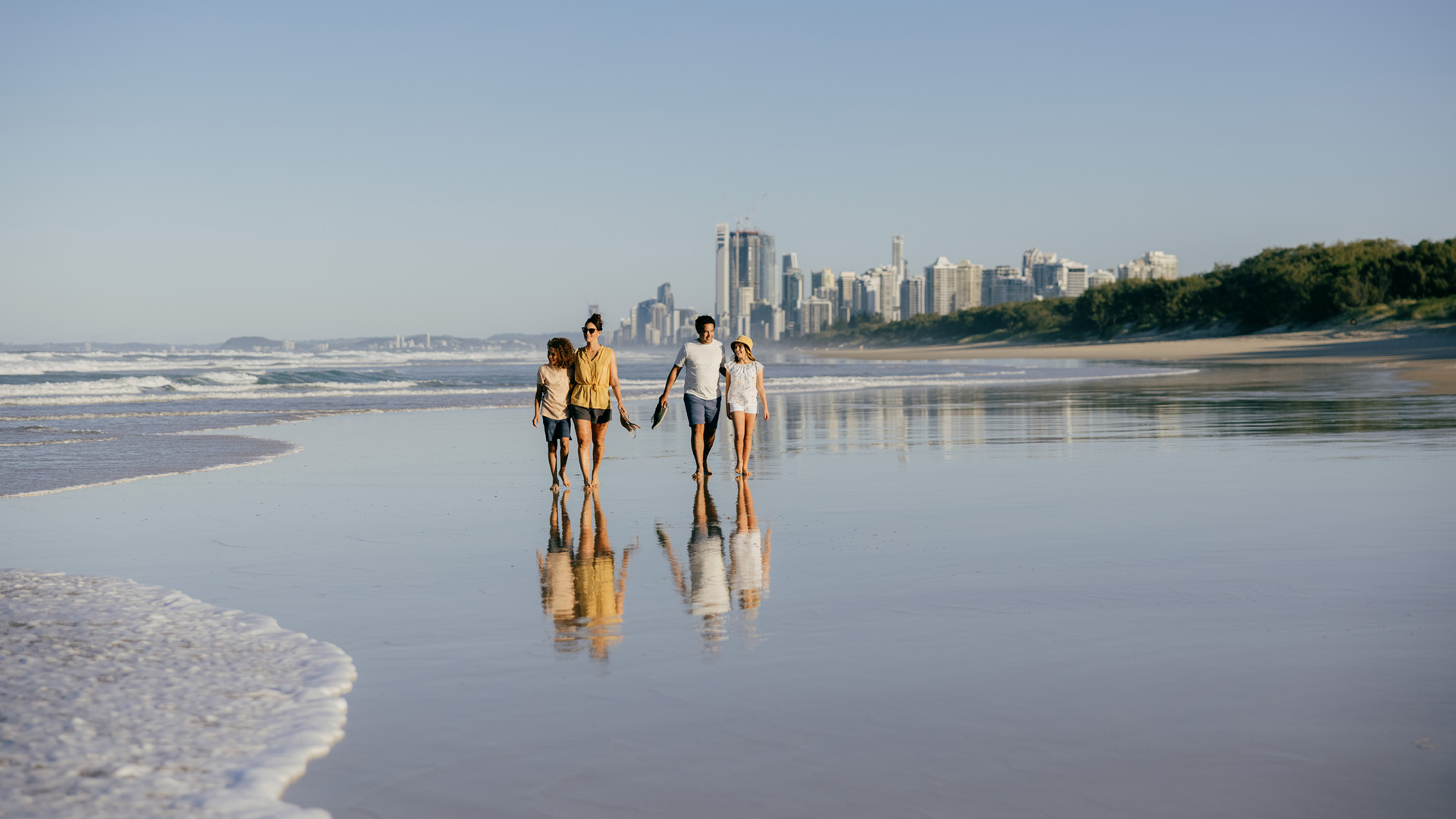 Four people walk hand in hand along a sandy beach by the ocean, with a city skyline visible in the background under a clear blue sky.