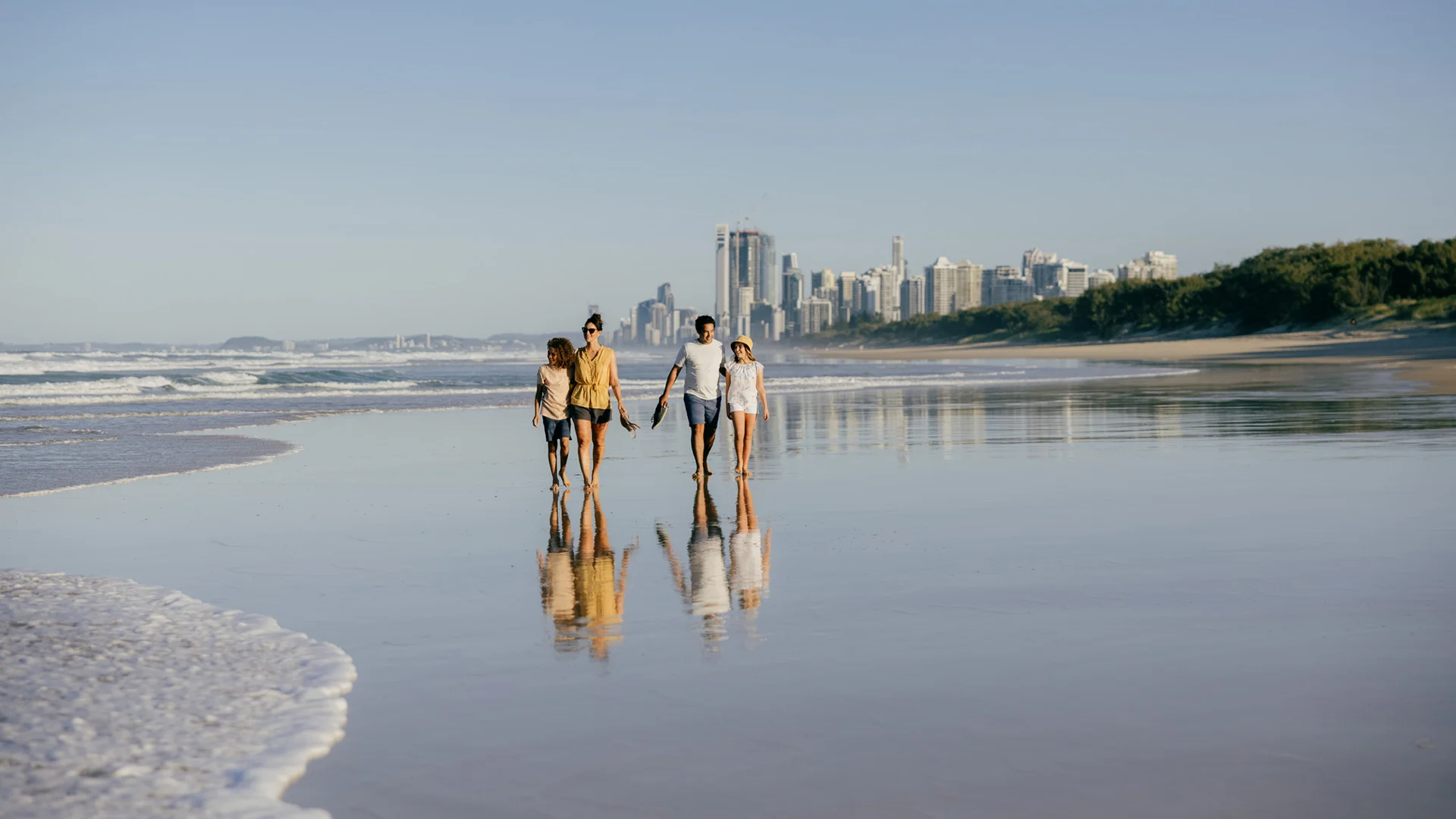 Four people walk hand in hand along a sandy beach by the ocean, with a city skyline visible in the background under a clear blue sky.