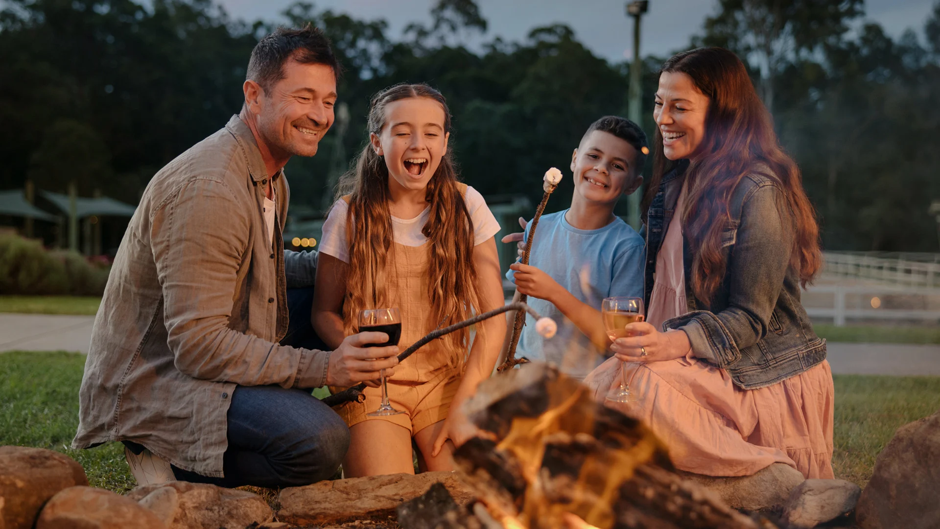 A family of four sits around a campfire outdoors, smiling and roasting marshmallows, with trees and buildings visible in the background.