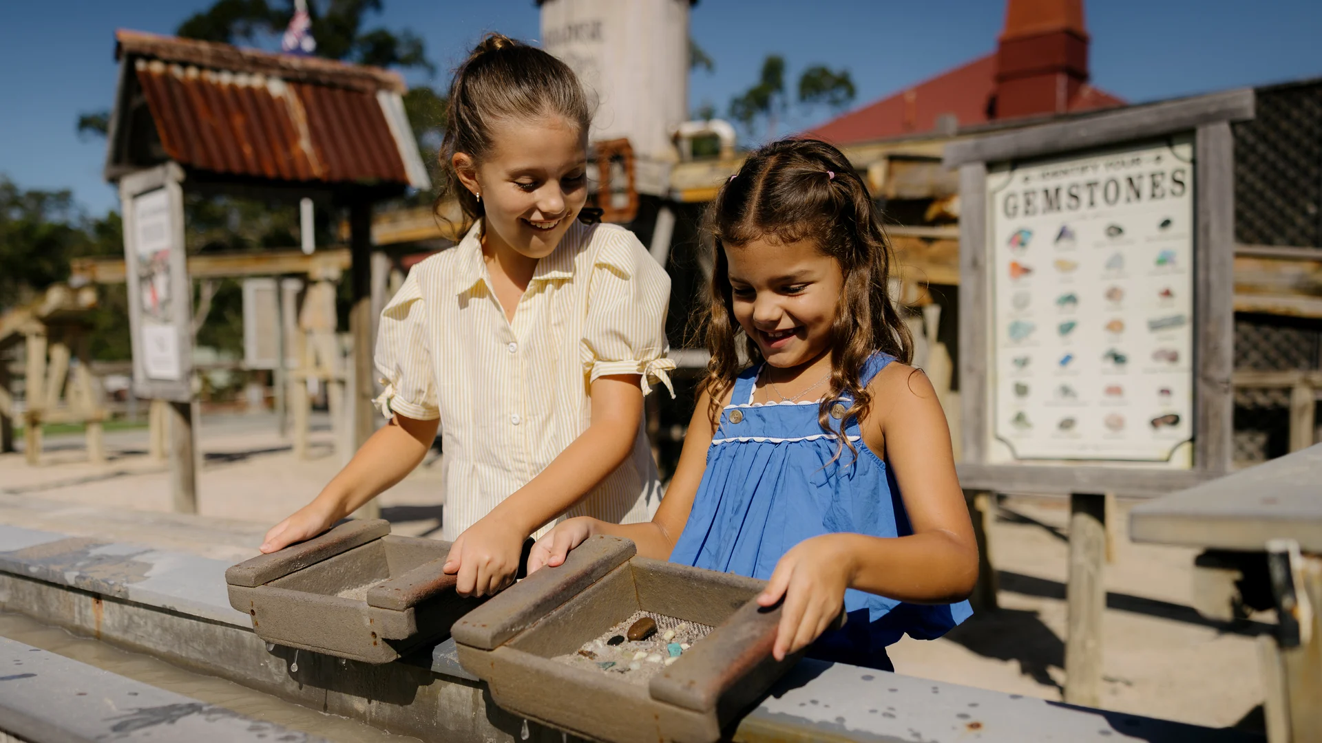 Two young girls smile as they sift through trays of rocks and water at an outdoor gemstone mining activity. A sign listing gemstones is visible behind them, and the setting resembles an old-fashioned mining town.