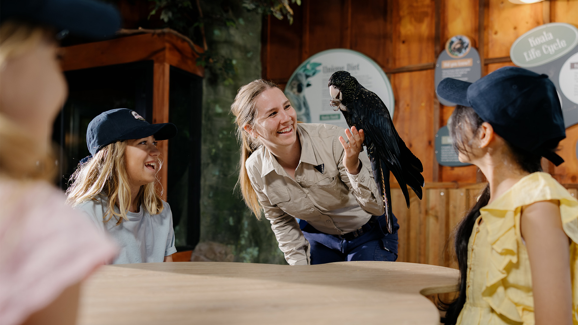 A woman holds a black parrot while smiling and interacting with three children wearing hats, gathered around a table in an indoor, nature-themed setting.