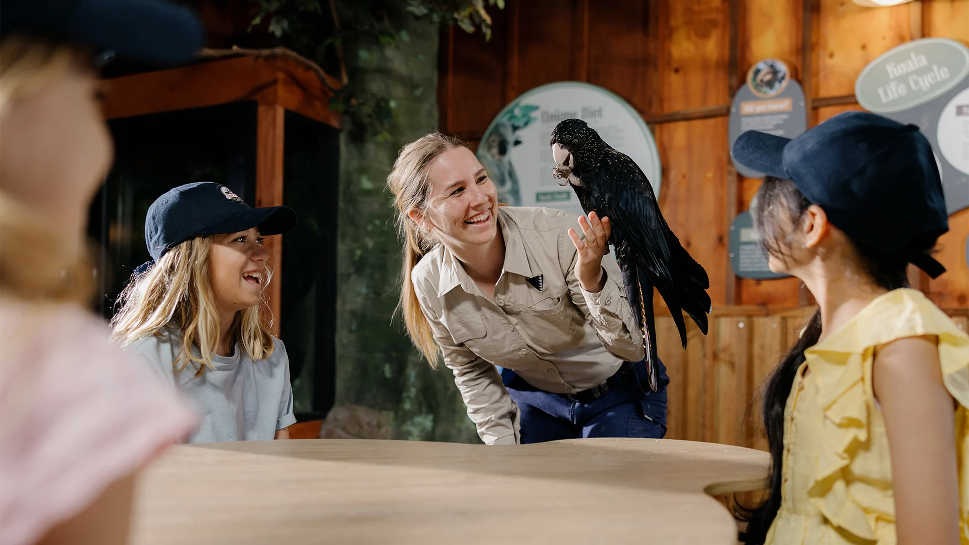 A woman holds a black parrot while smiling and interacting with three children wearing hats, gathered around a table in an indoor, nature-themed setting.