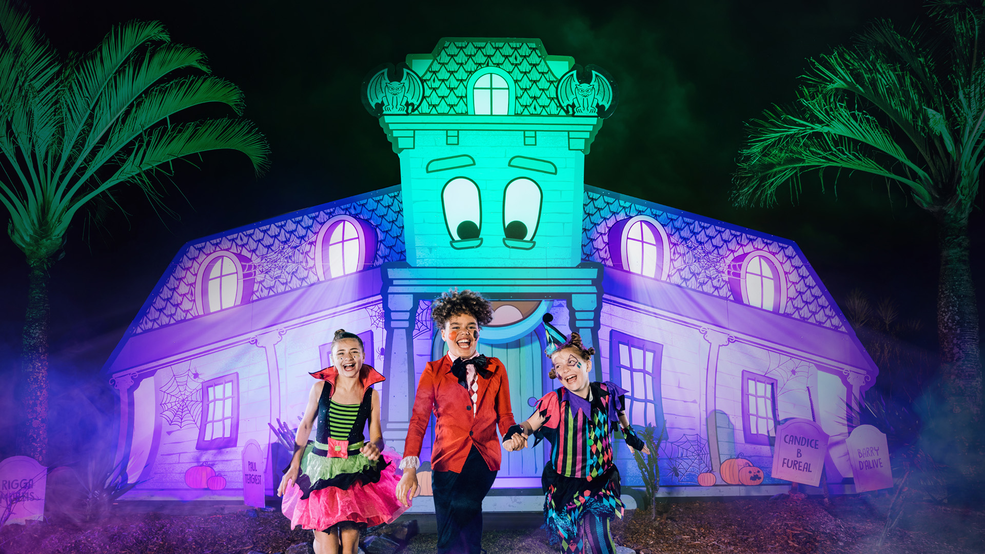 Three children in colorful Halloween costumes stand smiling in front of a brightly lit, cartoonish haunted house with large, animated eyes, surrounded by palm trees and Halloween decorations at night.