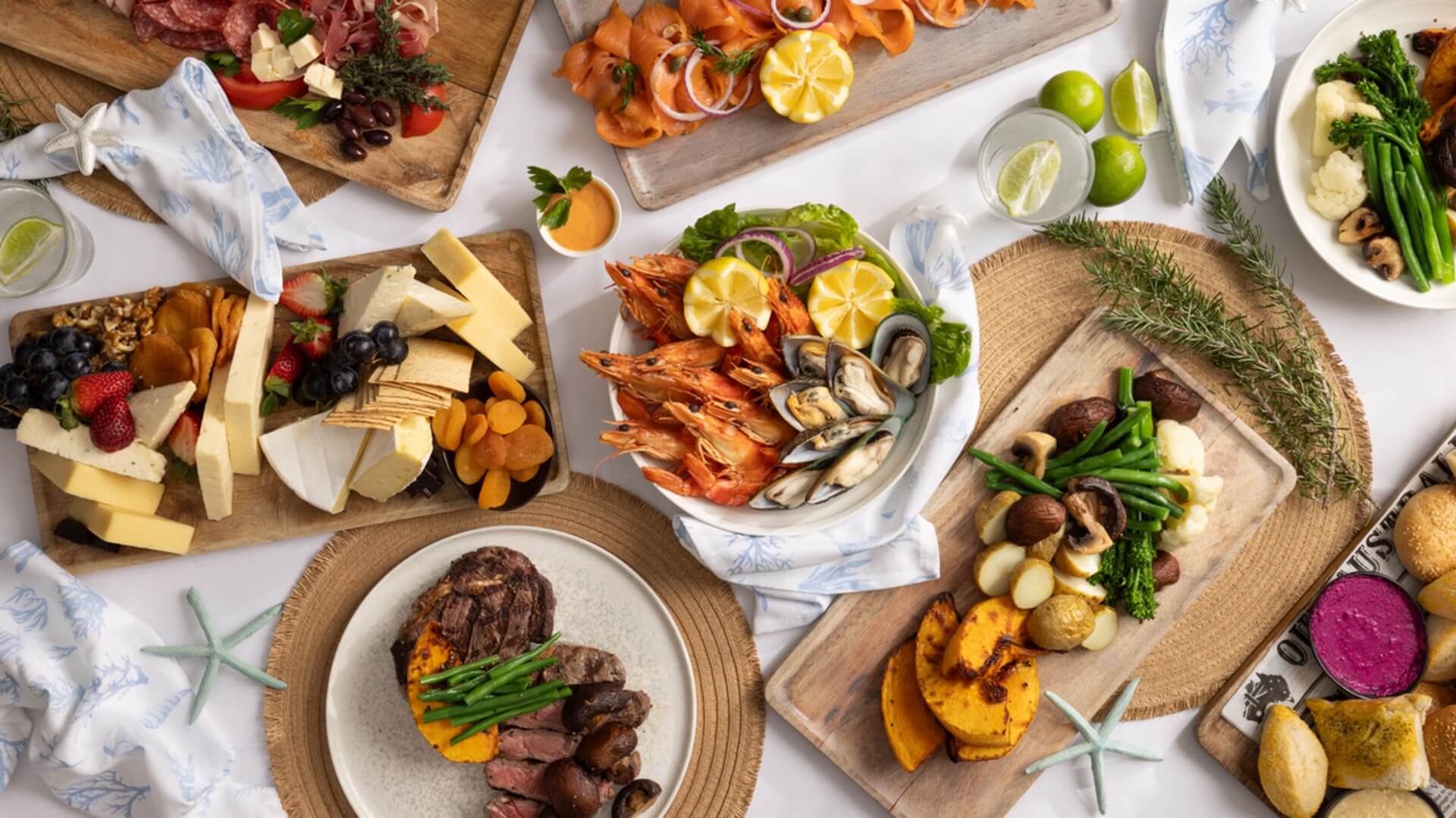 An assortment of dishes on a table, including cheese, seafood, meats, and vegetables, garnished with herbs and citrus slices. Drinks with lime are also present.
