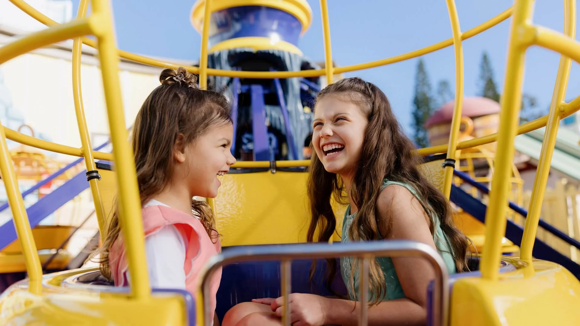 Two young girls with long hair sit smiling and laughing together on a bright yellow amusement park ride, enjoying a sunny day with colorful structures in the background.