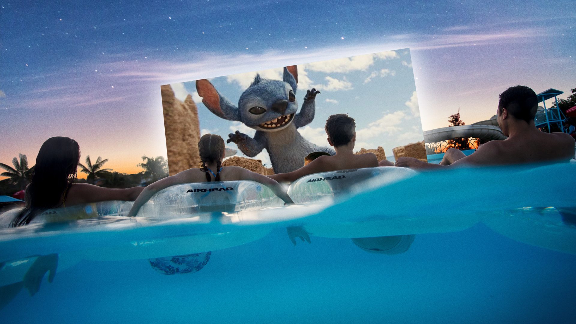 Four people relax in pool floats at night, watching an outdoor movie projected on a large screen above the water, showing a scene with an animated blue creature waving. Palm trees are visible in the background.