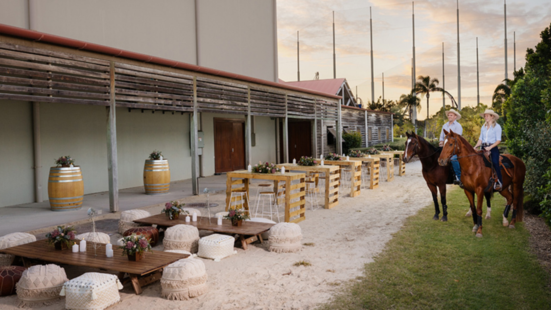 Two people in cowboy hats ride horses past a rustic outdoor seating area with wooden tables, stools, and barrels beside a large building at sunset.