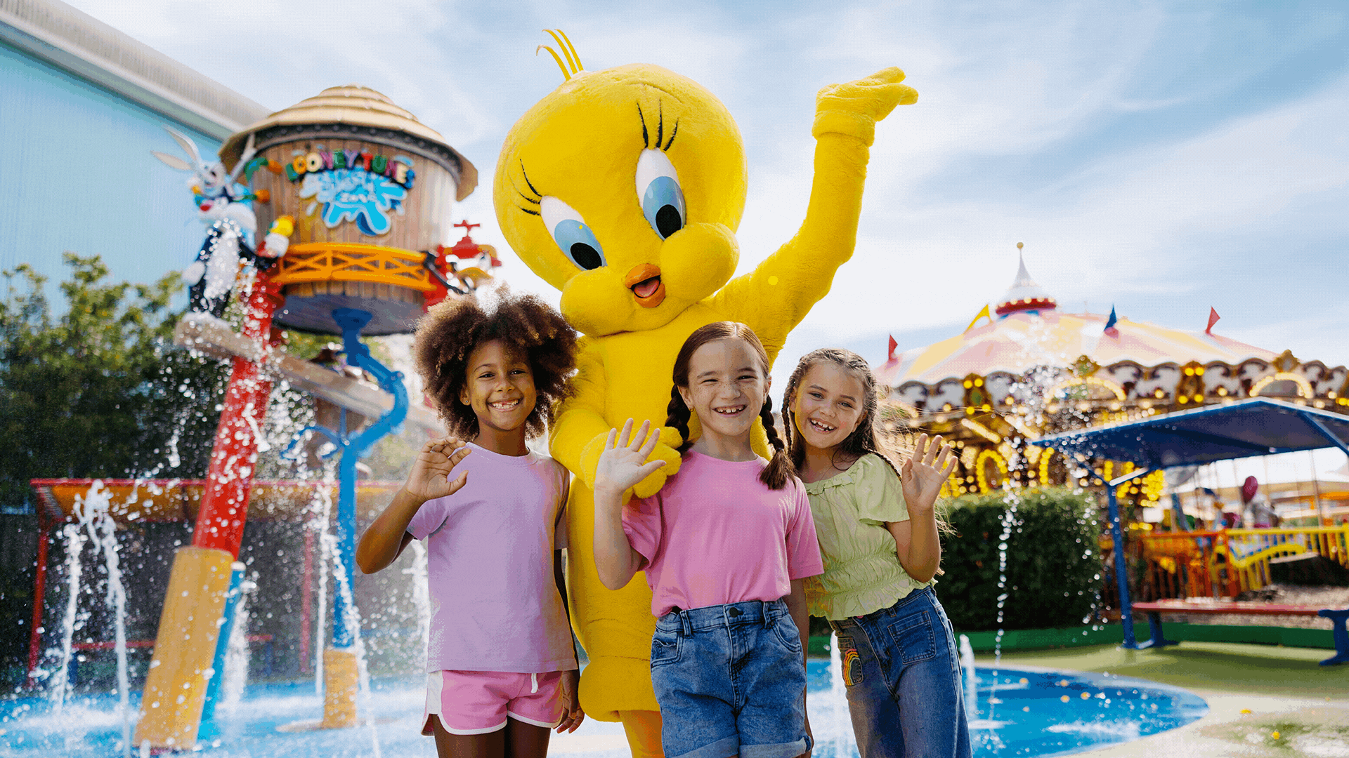 Three girls stand smiling and waving next to a person in a Tweety Bird costume at an amusement park, with a carousel and water play area in the background.