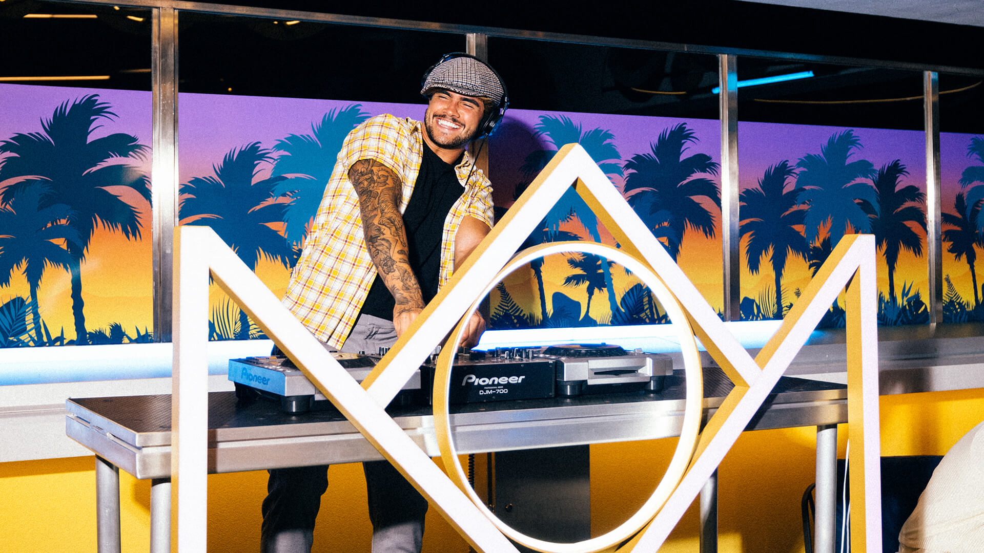 A smiling DJ in a checkered shirt and flat cap plays music at a turntable in front of a colorful backdrop with palm trees and a sunset. A geometric, crown-like frame decorates the DJ booth.