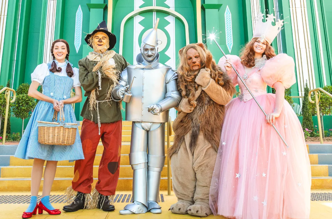 Five people in colorful costumes pose as Dorothy, the Scarecrow, Tin Man, Cowardly Lion, and Glinda the Good Witch from "The Wizard of Oz," standing on yellow steps in front of a bright green building.