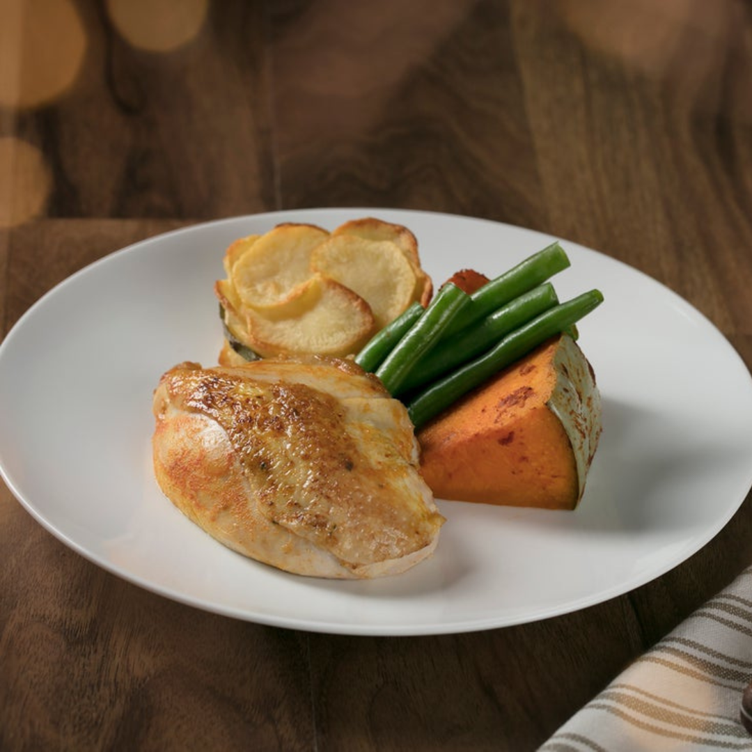A white plate with roasted chicken breast, a stack of potato slices, cooked green beans, and a large wedge of baked sweet potato, set on a wooden table with a striped napkin nearby.