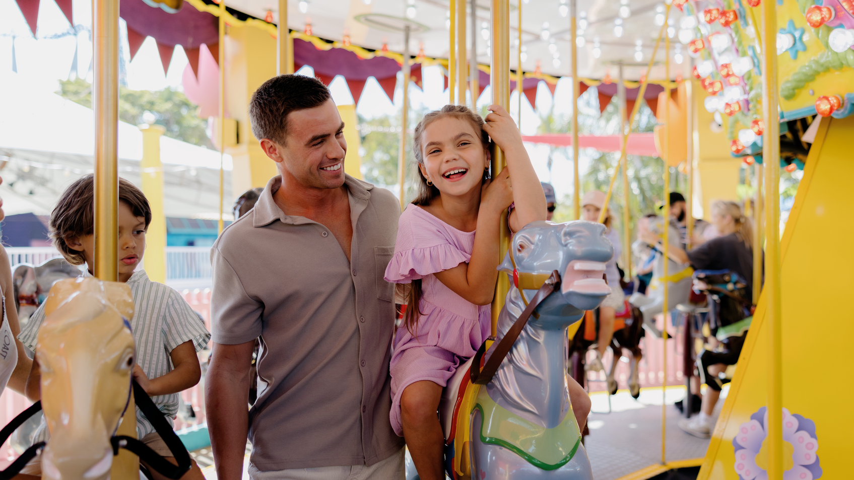 A man stands beside a smiling young girl riding a colorful carousel horse at an amusement park. Other children and adults are also enjoying the ride in the background.
