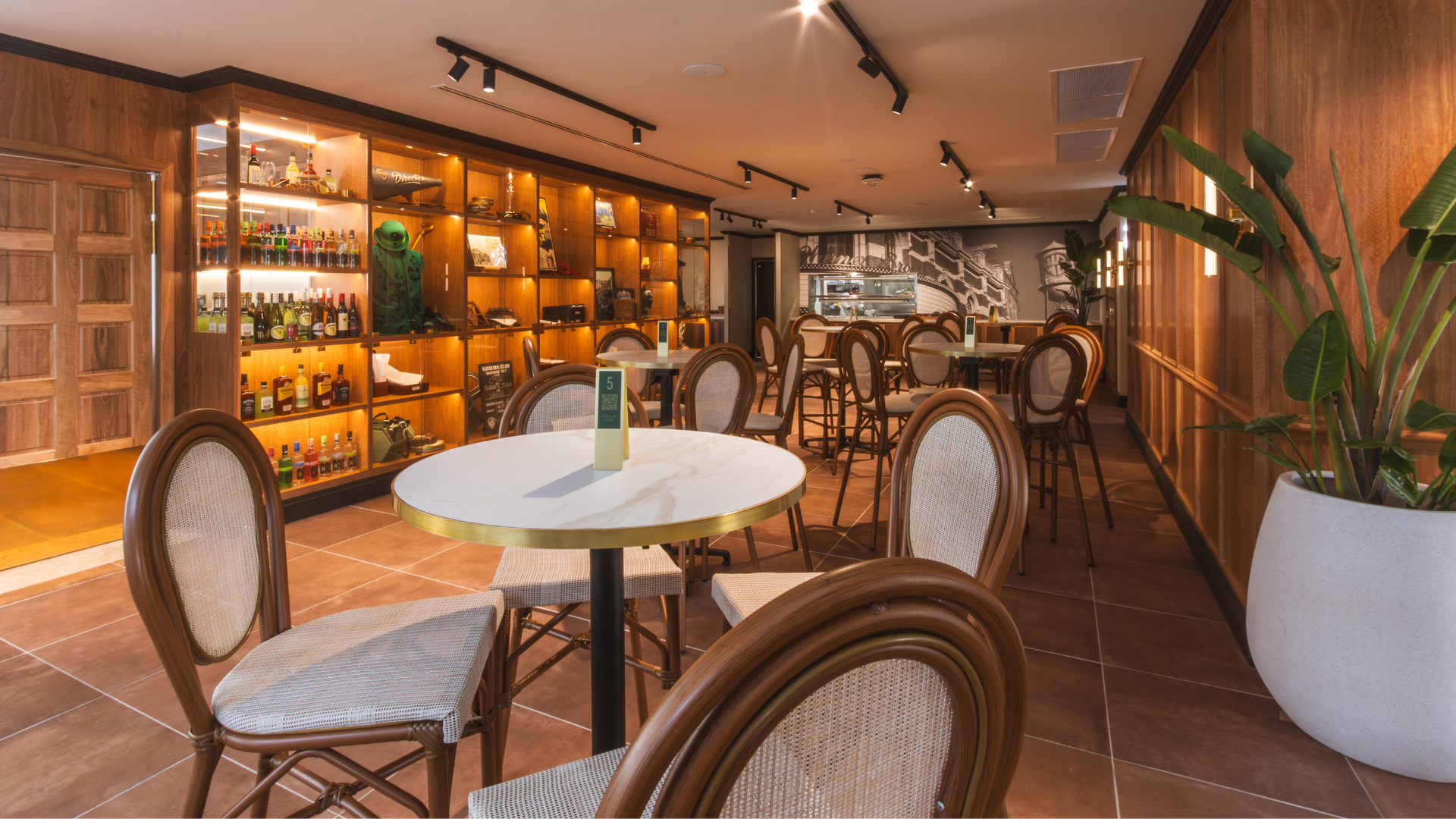 A cozy café interior with round tables, wicker chairs, wooden walls, shelves displaying bottles and decor, a large potted plant, and a counter in the background. Warm lighting creates a welcoming atmosphere.