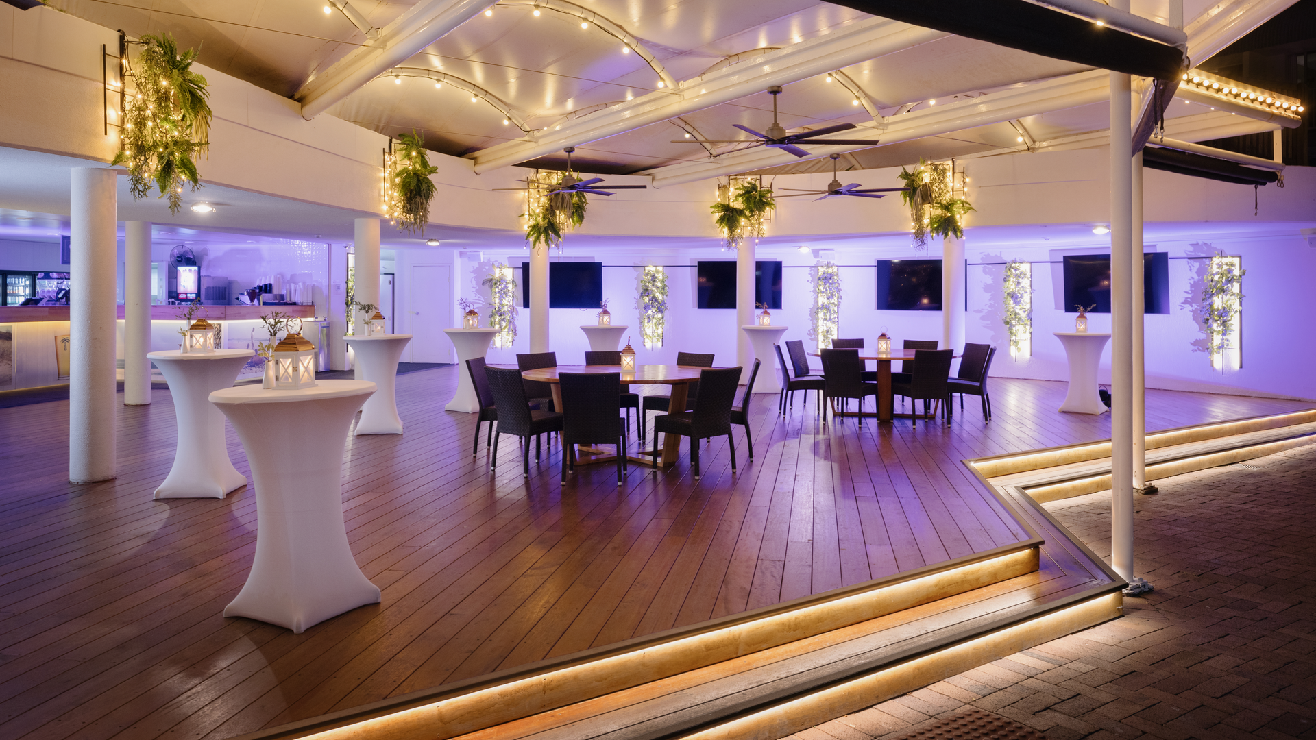 A modern, open-air event space with wooden floors, high tables with white covers, round tables with chairs, overhead string lights, hanging plants, and a softly lit bar area in the background.