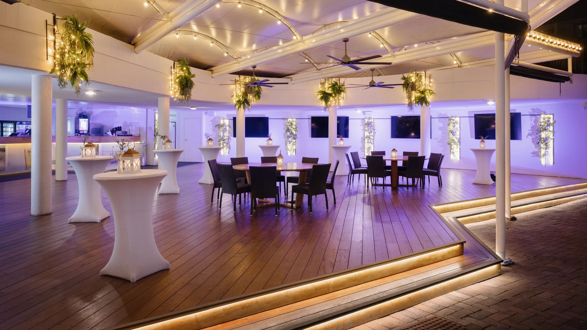 A modern, open-air event space with wooden floors, high tables with white covers, round tables with chairs, overhead string lights, hanging plants, and a softly lit bar area in the background.