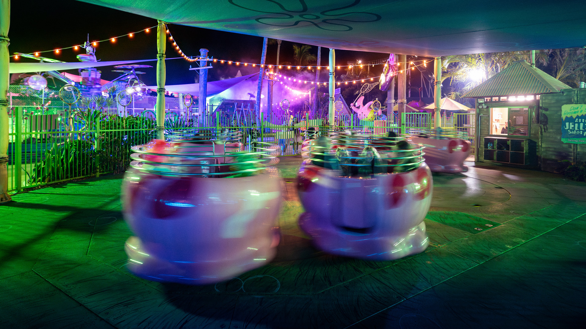 Colorful spinning teacup ride at an amusement park at night, with bright lights and blurred motion from the spinning cups, under a canopy with festive string lights in the background.