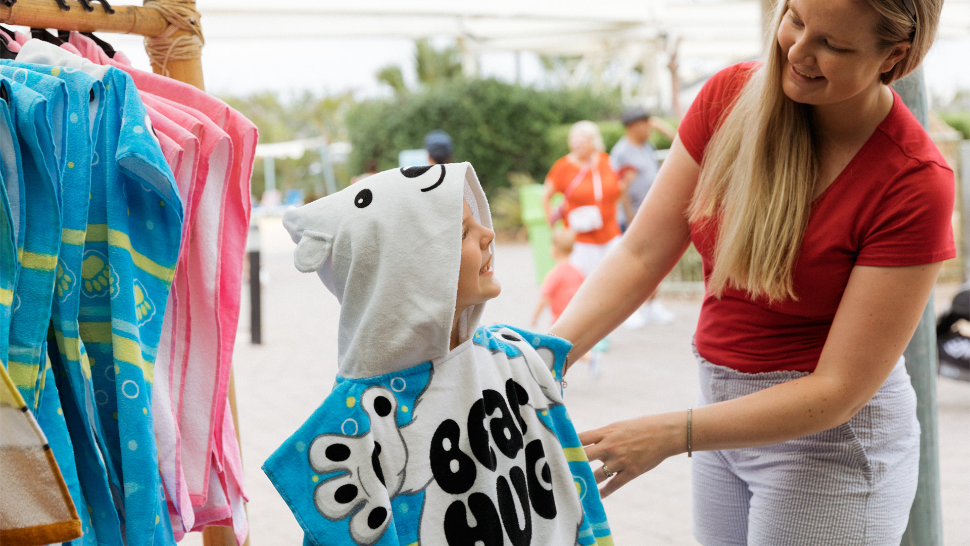 A woman helps a child try on a hooded towel with a bear design and the words "Bear Hug" at an outdoor shop.