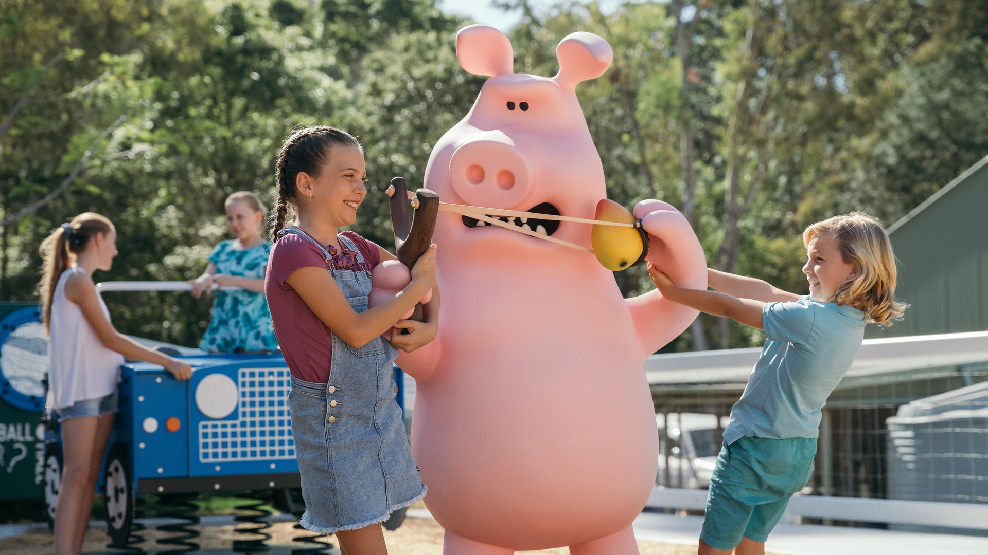 Two children play with a large pink pig mascot outdoors, smiling and using a stretchy toy, while adults watch in the background.