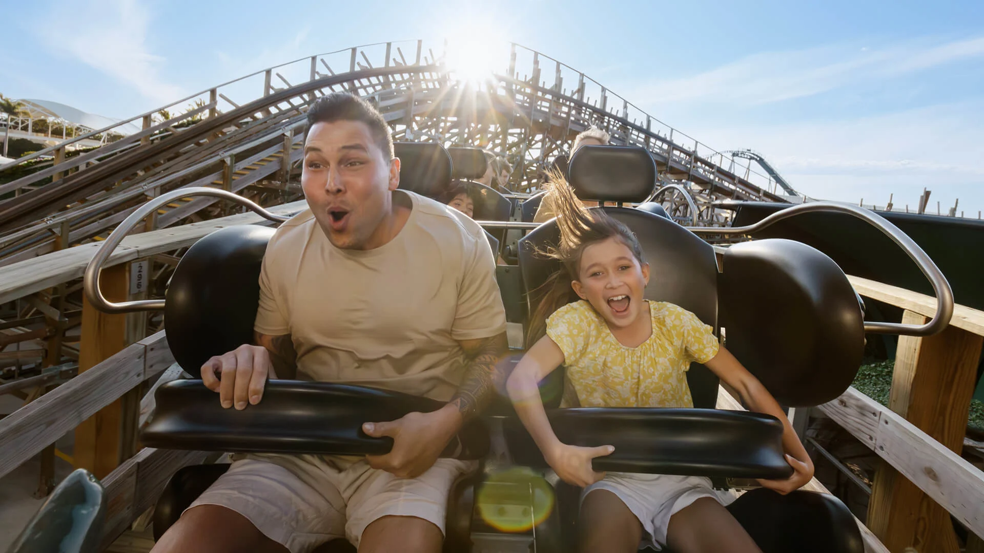 An adult and a child ride a wooden roller coaster, gripping the safety bars, with excited and surprised expressions as the sun shines in the background.