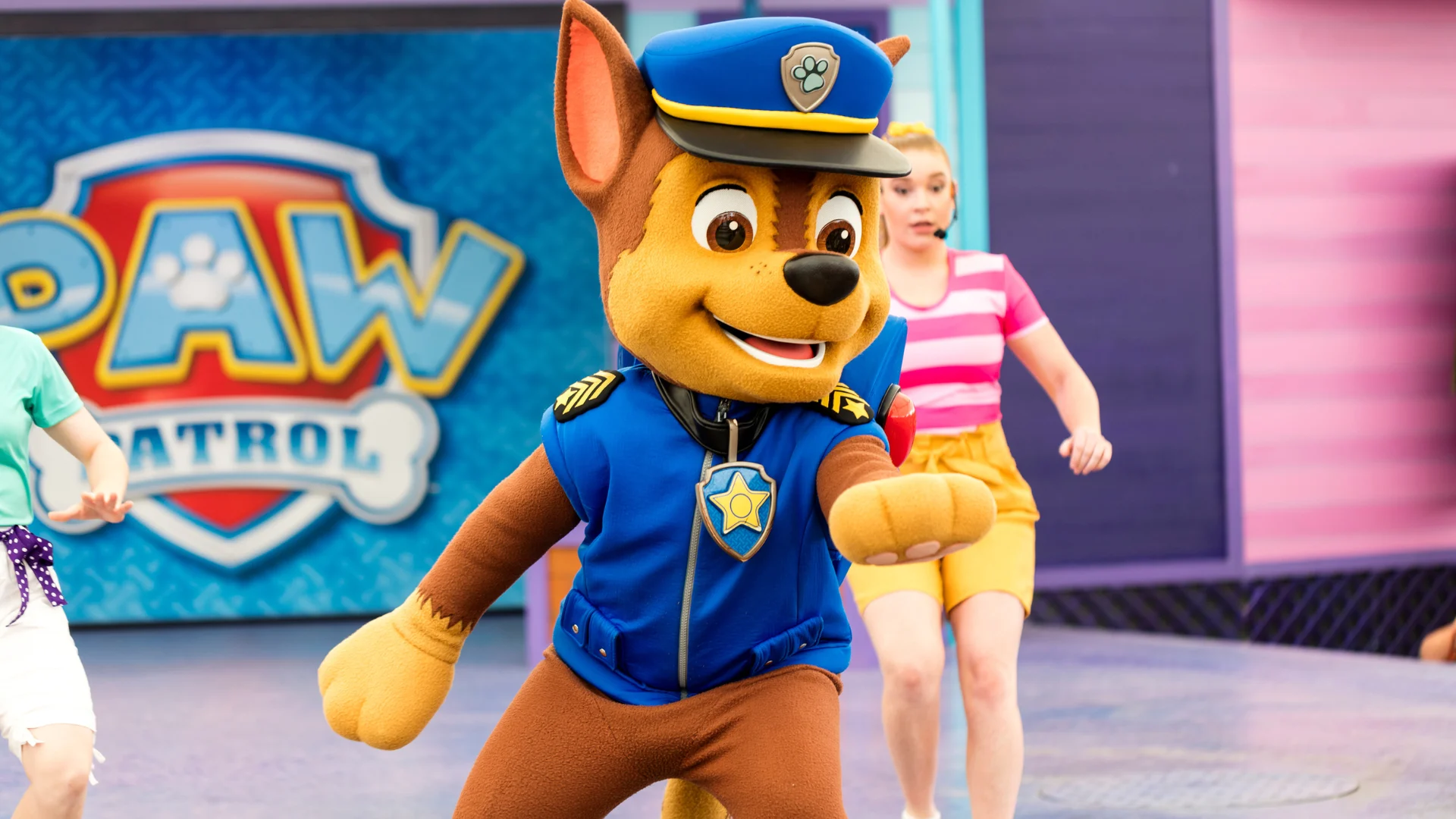 A person in a Chase costume from PAW Patrol performs on stage with two dancers, one wearing a pink striped shirt and yellow shorts. The PAW Patrol logo is visible in the background.