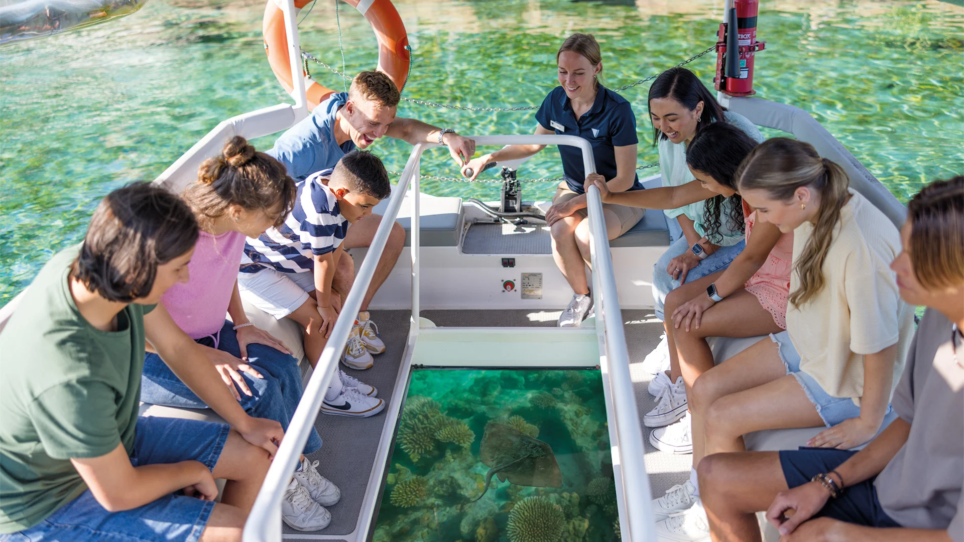 A group of people sit in a glass-bottom boat, looking down at coral and marine life through the glass panel. The boat is on clear, shallow water.