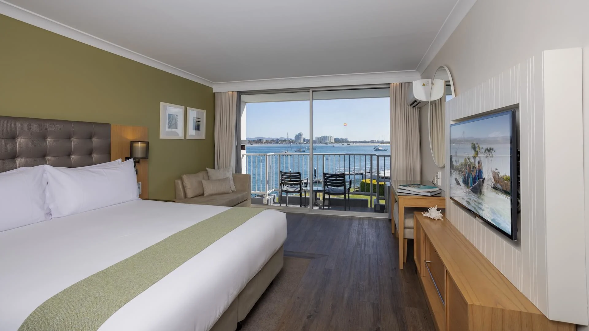 Hotel room with a large bed, a wall-mounted TV, a sofa, and a desk. A sliding glass door opens to a balcony with a view of the waterfront and city skyline.